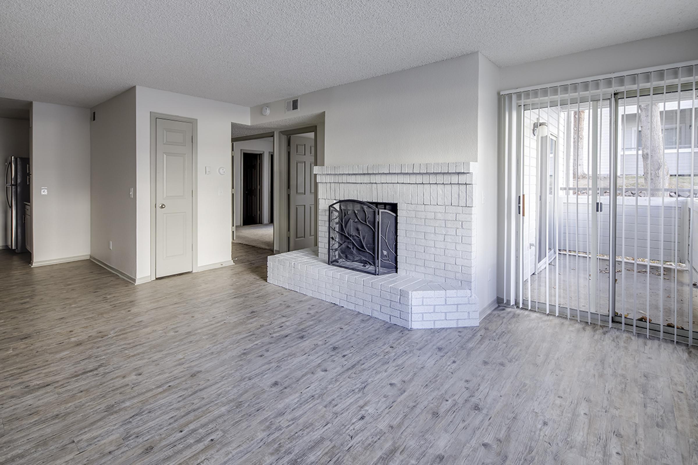 Spacious living area featuring light-colored laminate flooring, a white brick fireplace, and large windows with sliding glass doors leading to an outdoor space. Neutral wall colors create a bright, inviting atmosphere. Doorways lead to additional rooms, adding to the layout's openness.