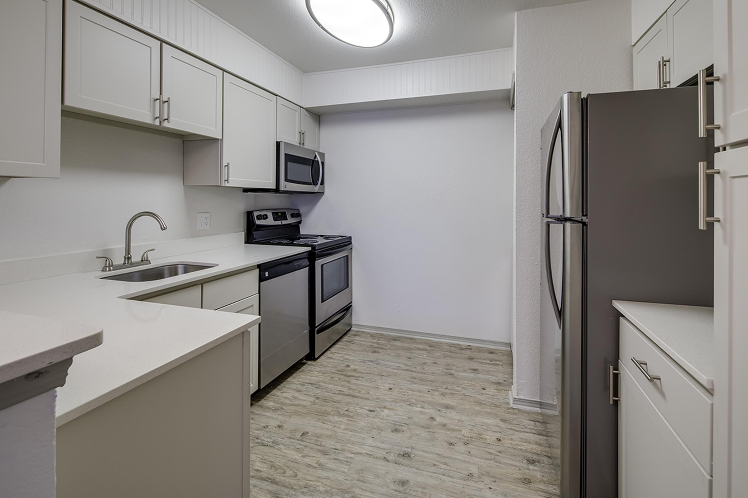 Modern kitchen featuring light-colored cabinetry, stainless steel appliances including a refrigerator, stove, and microwave. The countertop is white, with a sink positioned beneath a window. The flooring is light wood, and the walls are painted white, creating a bright and airy atmosphere.