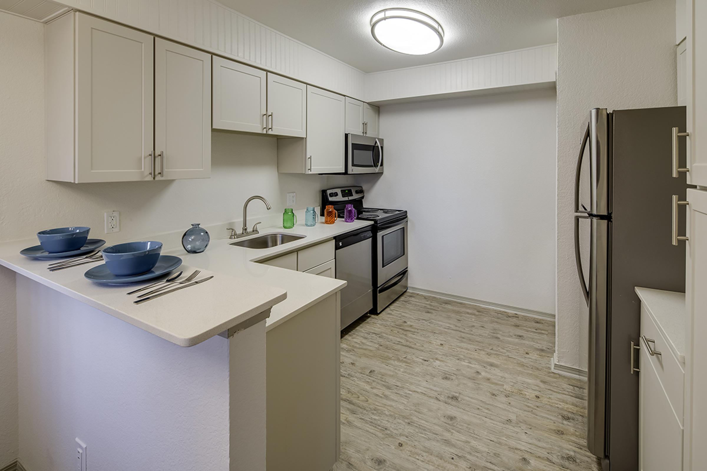 Modern kitchen featuring light-colored cabinets, stainless steel appliances, and a countertop with dining plates and utensils. The space includes a refrigerator, oven, sink, and ample natural light, creating a clean and inviting atmosphere. Durable wooden flooring complements the design.
