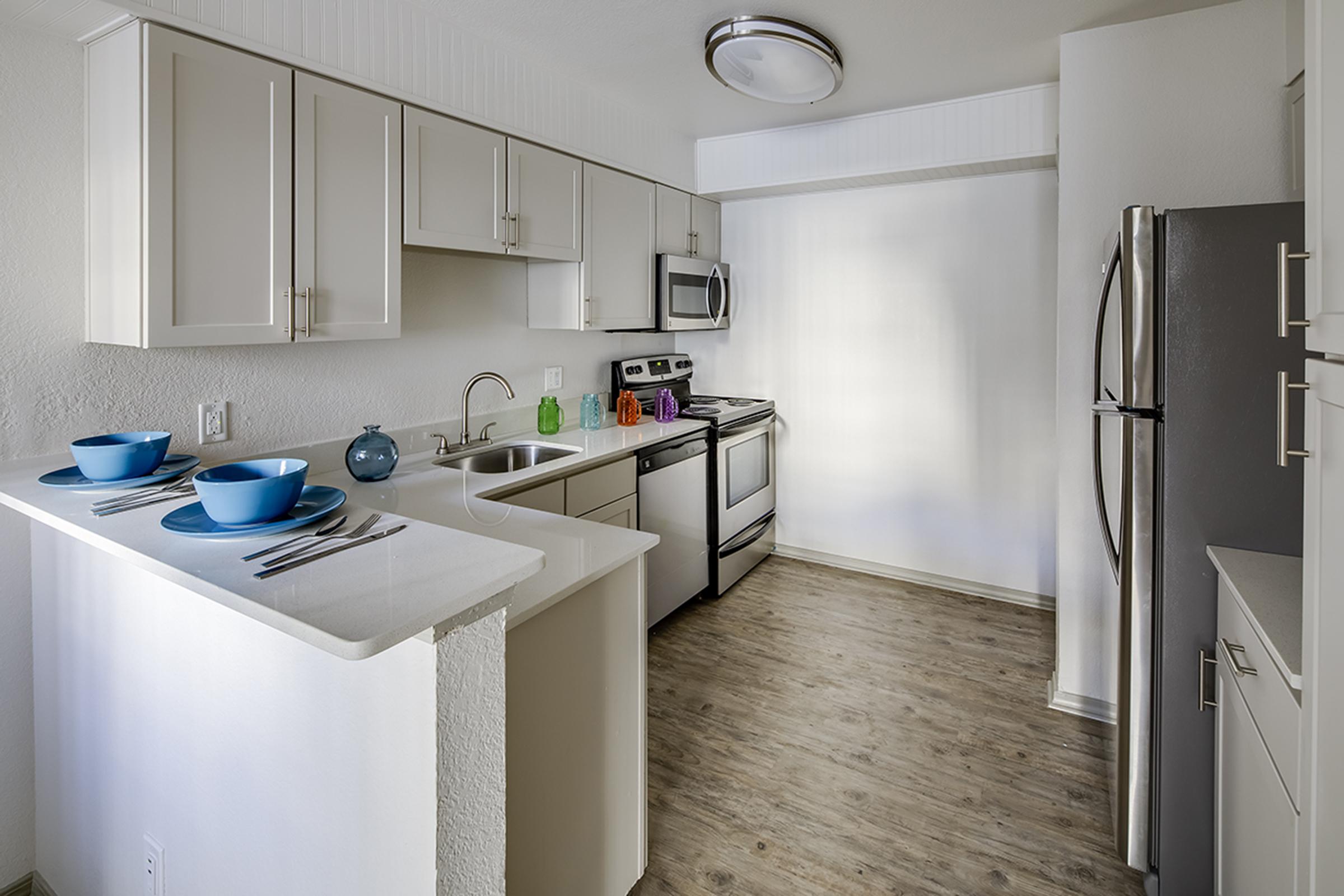 Modern kitchen with light-colored cabinets and stainless steel appliances. The countertop features two blue bowls, silver cutlery, and a small decorative vase. There's a spacious feel created by natural light, and the flooring is a warm wood-tone laminate.