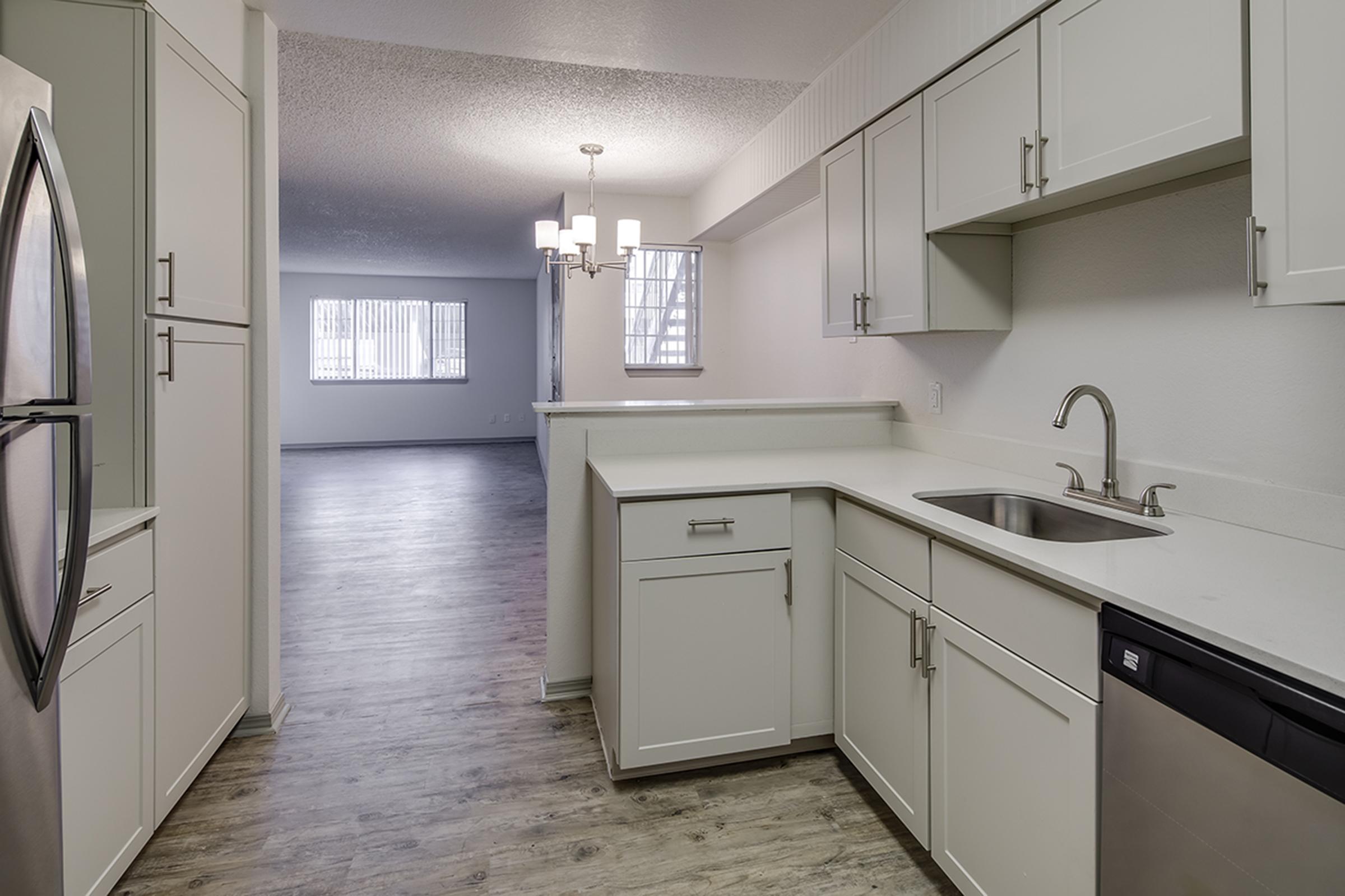 A modern kitchen featuring white cabinets, a stainless steel refrigerator, and a sink with a chrome faucet. The kitchen opens into a spacious living area with large windows allowing natural light, and light-colored flooring throughout. A dining area with a chandelier is visible.
