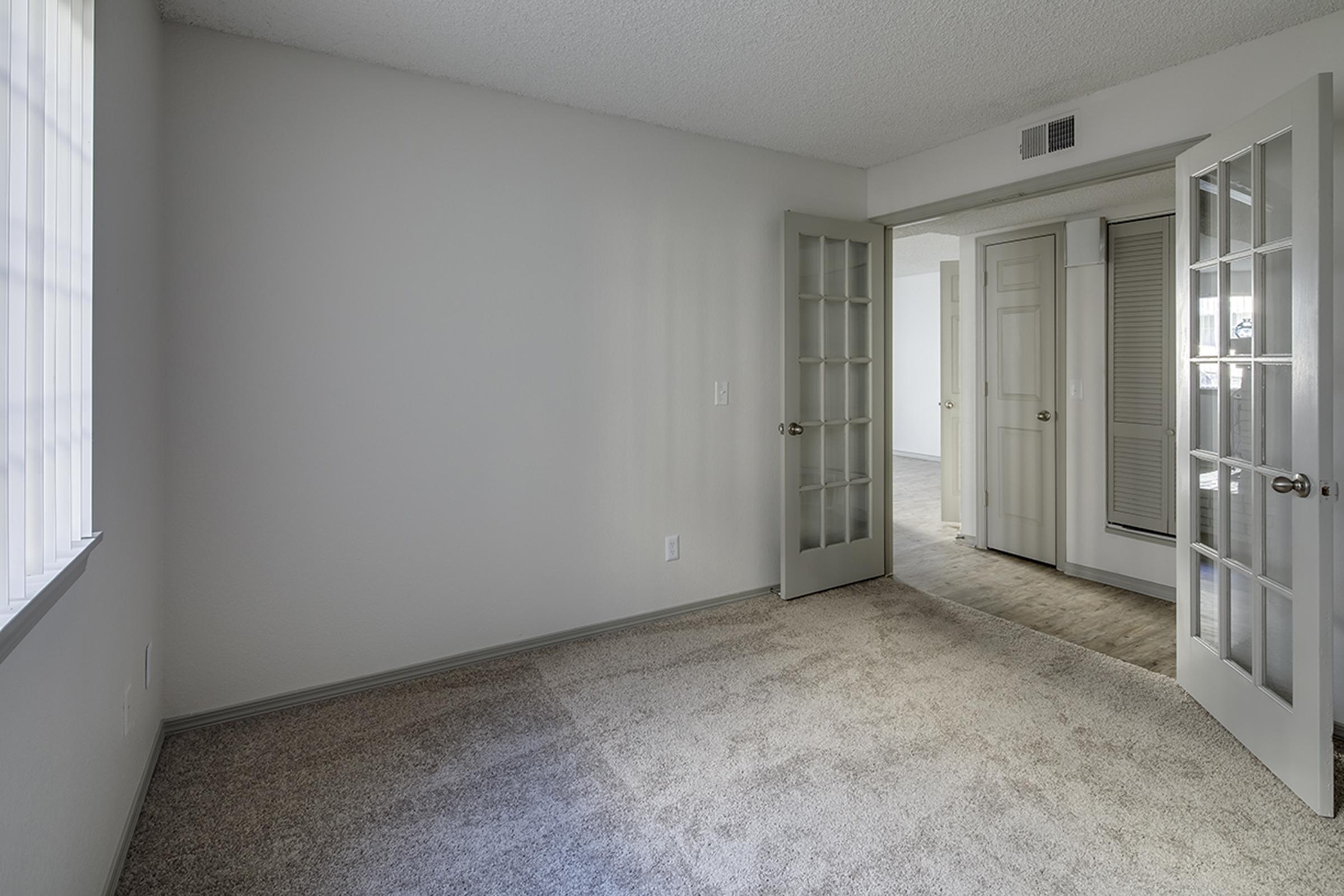 A spacious, empty room with light gray walls and carpet. There is a large window allowing natural light to fill the space. Two doors with glass panels open to a hallway leading to another room, which has neutral-colored walls and flooring. The overall ambiance is bright and inviting.
