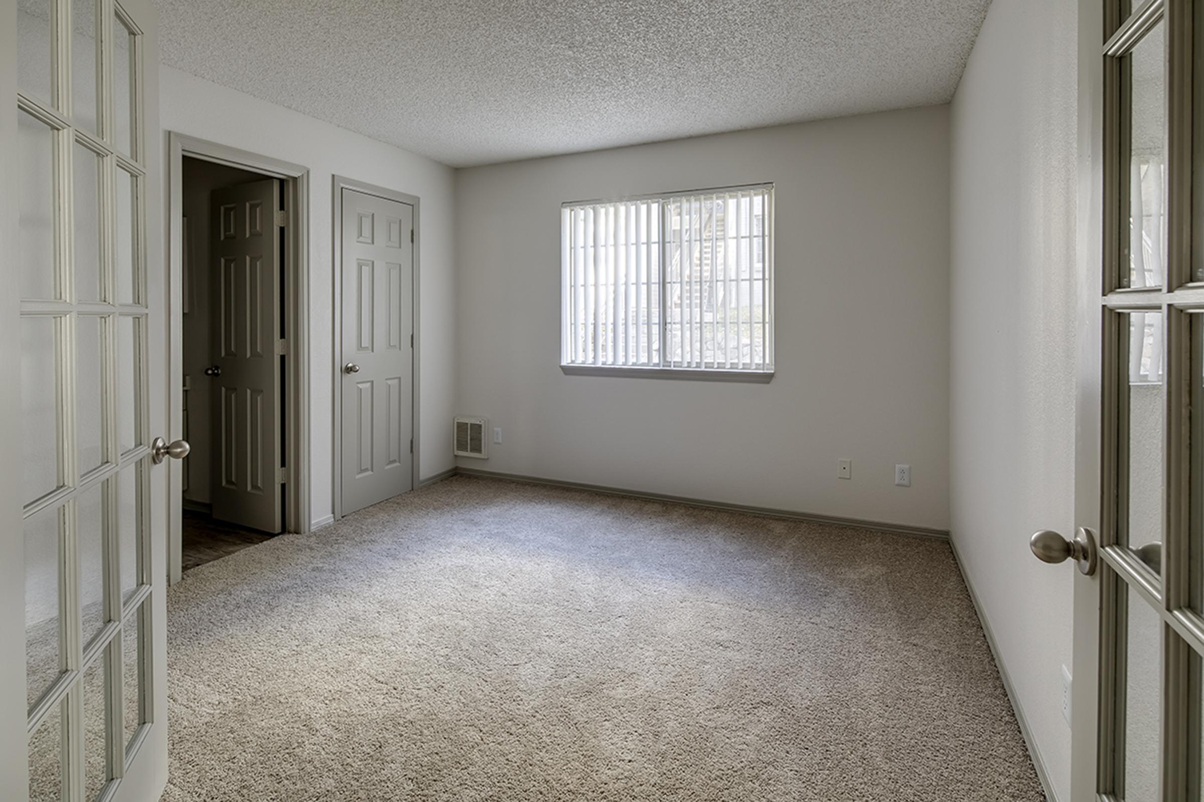 A vacant room with beige carpet and light-colored walls. It features two doors, one leading to another room and the other to a hallway. A window on the left lets in natural light, casting soft shadows on the floor. The space is clean and minimalistic, ideal for personal customization.
