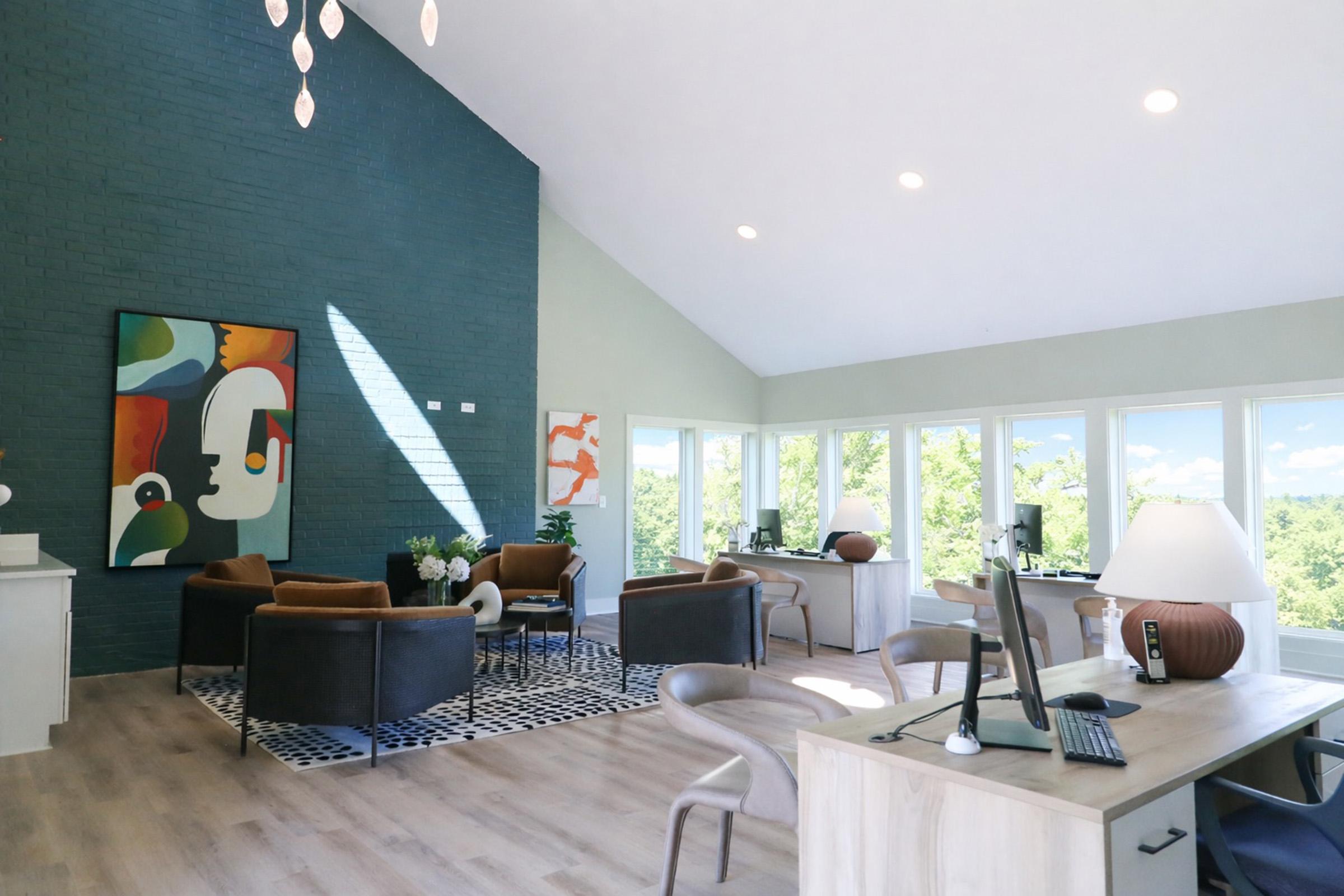 Bright and modern interior of a spacious room featuring large windows, contemporary furniture, and stylish decor. There are two desks, comfortable seating arrangements, and colorful artwork on the walls, creating an inviting atmosphere with plenty of natural light and greenery visible outside.