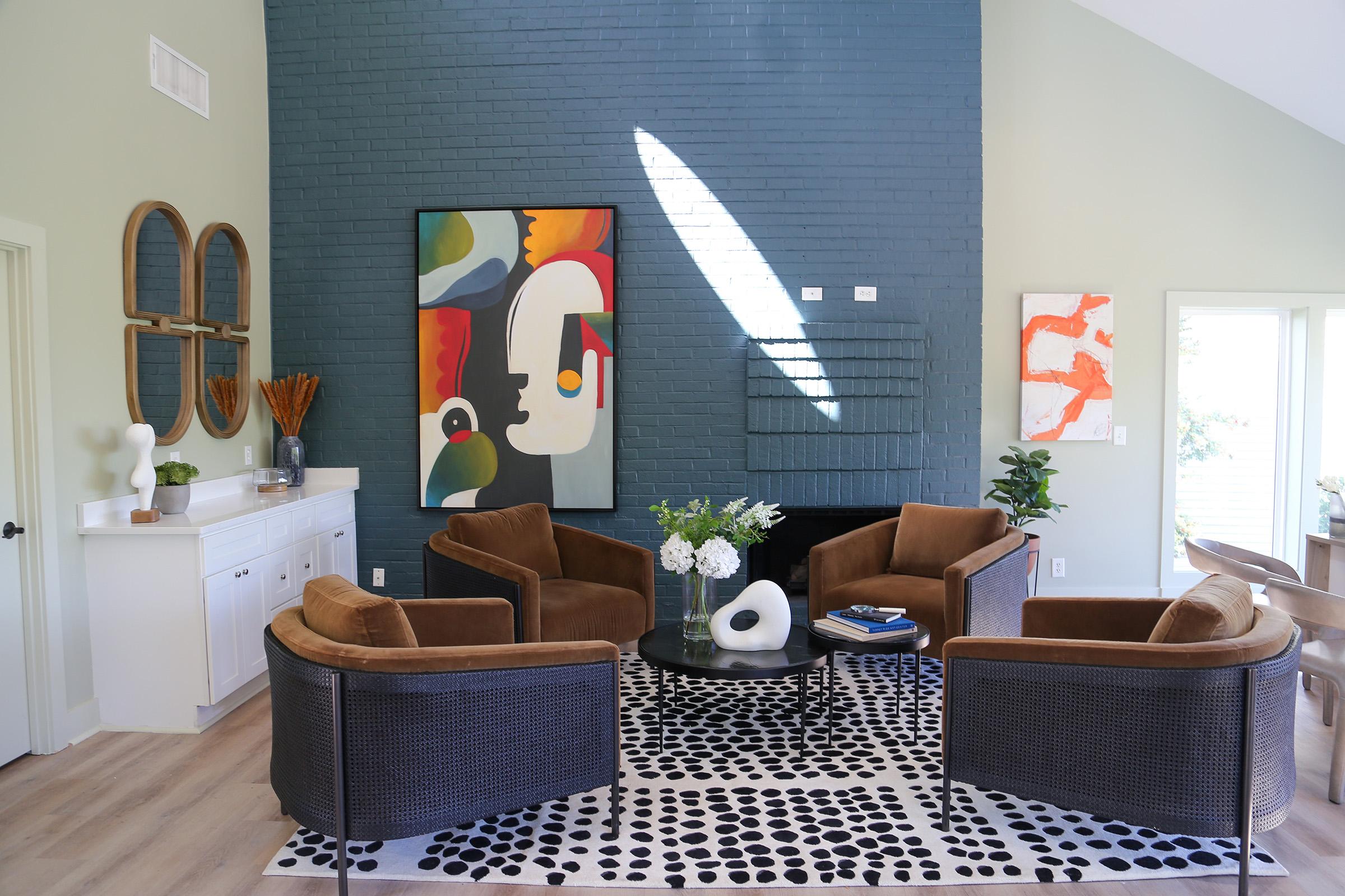A modern living room featuring two brown armchairs and a round glass coffee table on a black and white patterned rug. The walls are painted light green and dark blue, with abstract artwork hanging on the walls. A white sideboard holds decorative items and a vase with flowers, complemented by natural light from nearby windows.