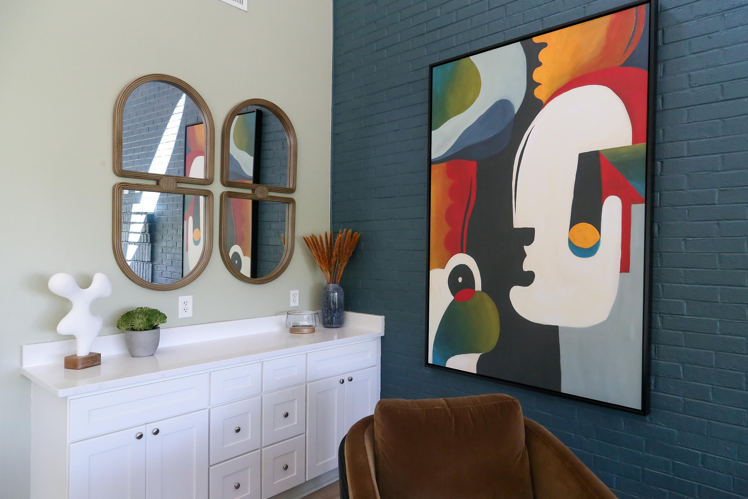 A modern living space featuring a white console table with drawers, three round mirrors on the wall, and a large abstract painting of a face. A brown chair and a decorative vase with dried plants add warmth to the room, while a small green plant sits on the table. The walls are a mix of light green and dark blue.