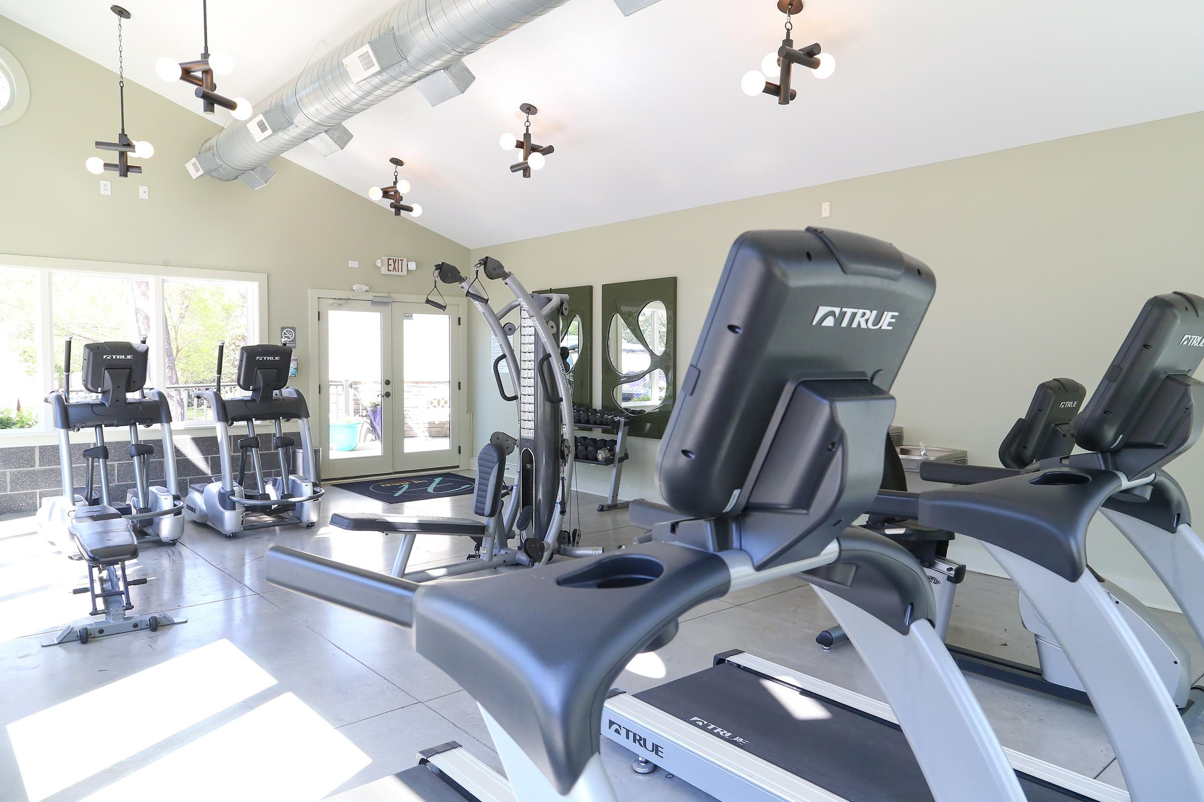 A modern fitness room featuring various exercise equipment including treadmills and elliptical machines, with large windows allowing natural light to enter. The interior has a sleek, contemporary design with light-colored walls and decorative light fixtures.