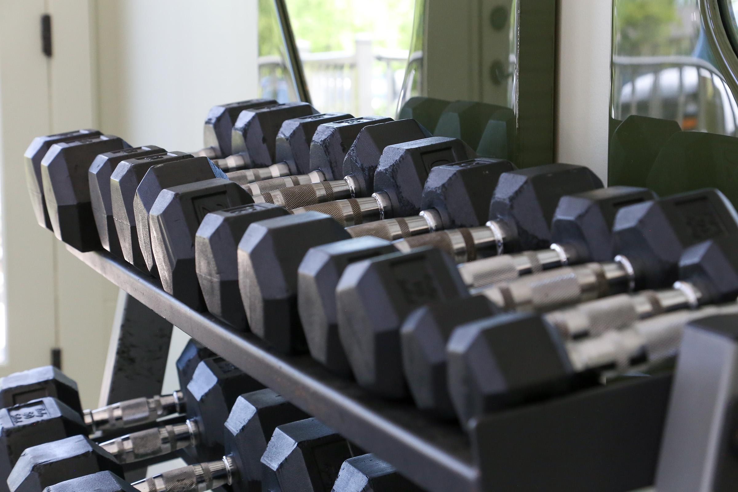 A rack of hexagonal black dumbbells arranged neatly in a gym setting. The dumbbells vary in weight and are positioned on a multi-tiered metal stand, with a mirror reflecting the equipment in the background, creating a functional fitness atmosphere.