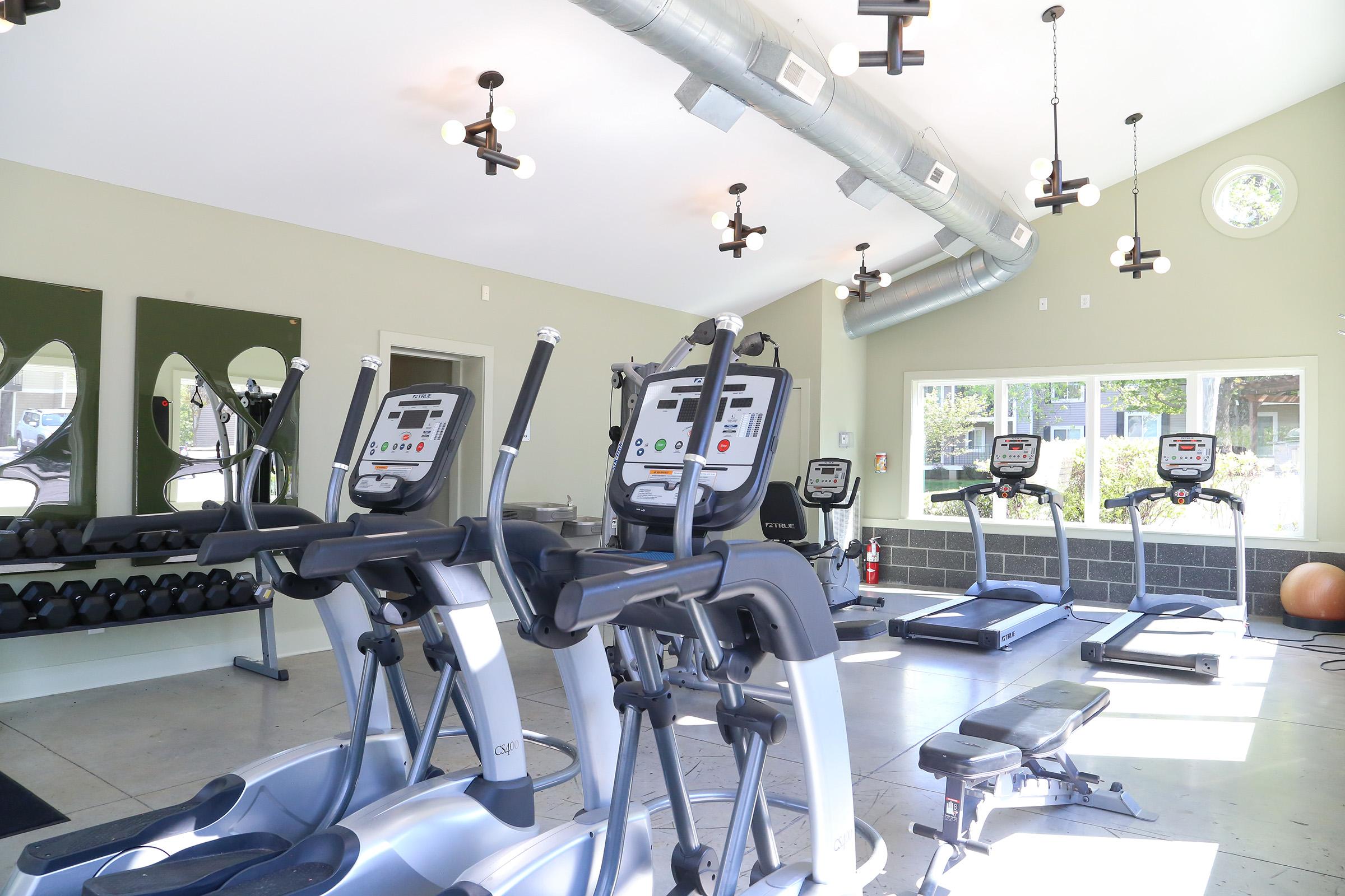 A modern gym interior featuring elliptical machines, treadmills, and weight benches. The space has large windows for natural light, sleek design with ventilation ducts, and various fitness equipment arranged neatly.