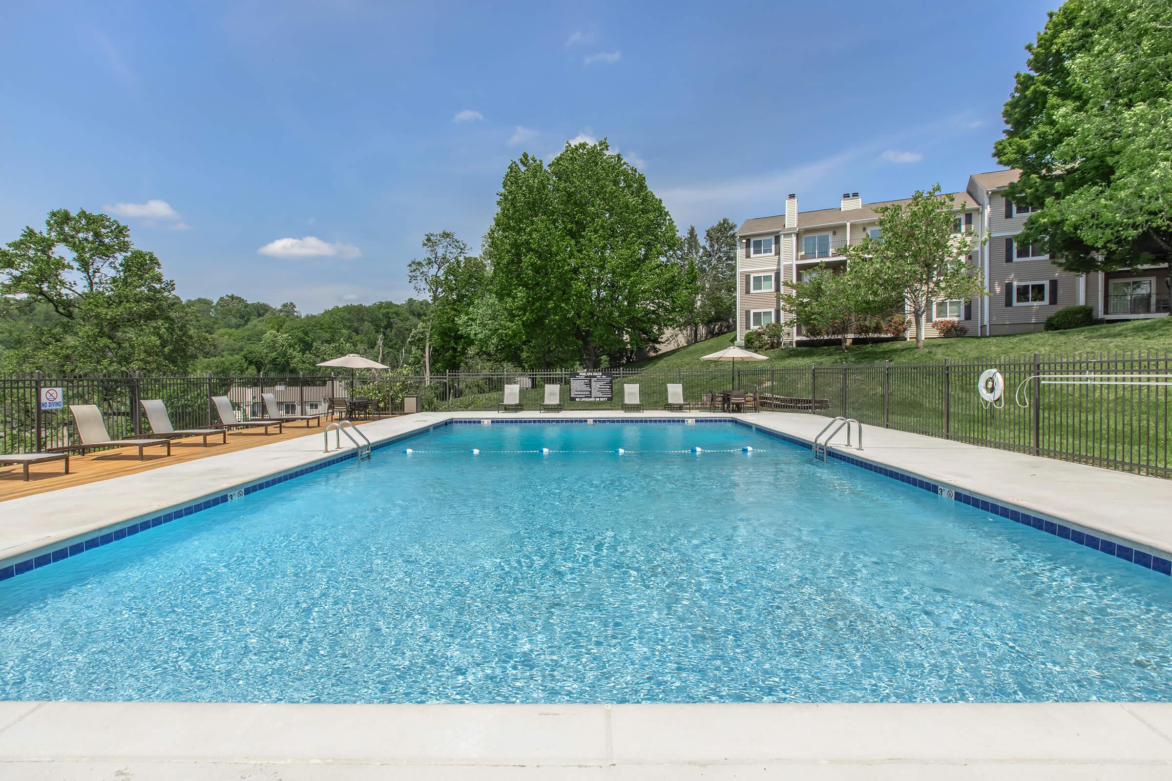 Swimming pool surrounded by a grassy area and residential buildings. The pool features clear blue water, lounge chairs on one side, and shaded areas with umbrellas. Lush green trees provide a scenic backdrop under a clear blue sky with a few clouds.
