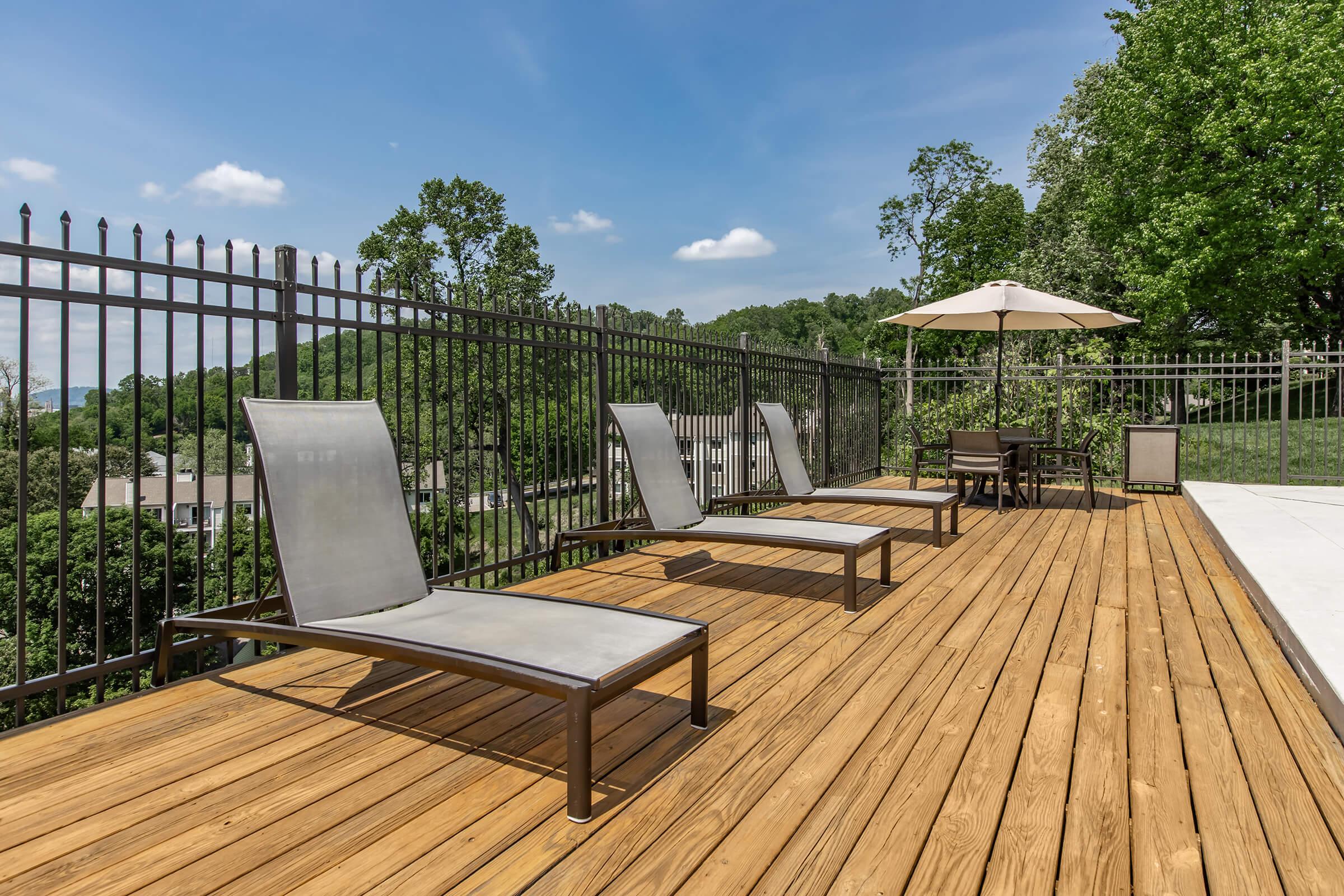 A scenic outdoor deck featuring two modern lounge chairs positioned on a wooden surface. A patio table and umbrella are visible, along with a green landscape in the background, including trees and a clear blue sky with a few clouds. The area is enclosed by a black metal fence.