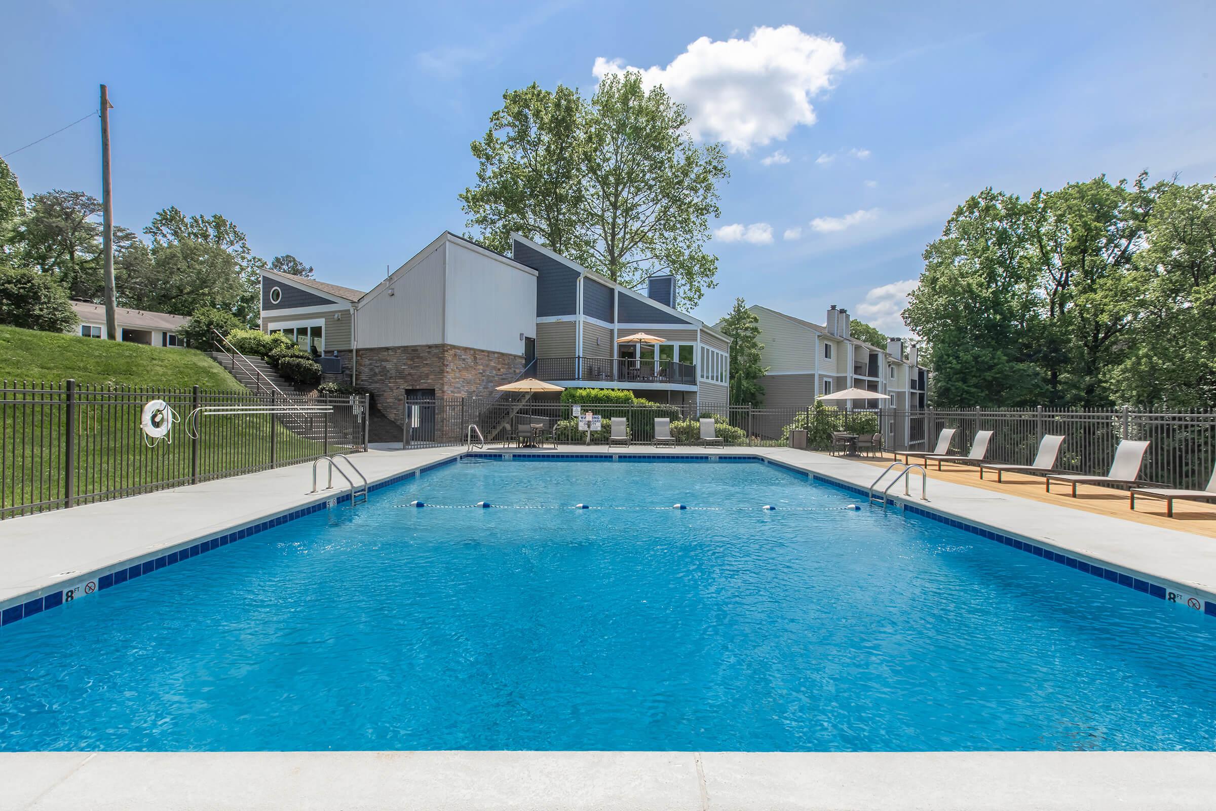 A clear blue swimming pool surrounded by a fenced patio area. Lounge chairs are positioned beside the pool, and a large umbrella provides shade. In the background, a modern building with trees and greenery can be seen under a bright blue sky with a few clouds.