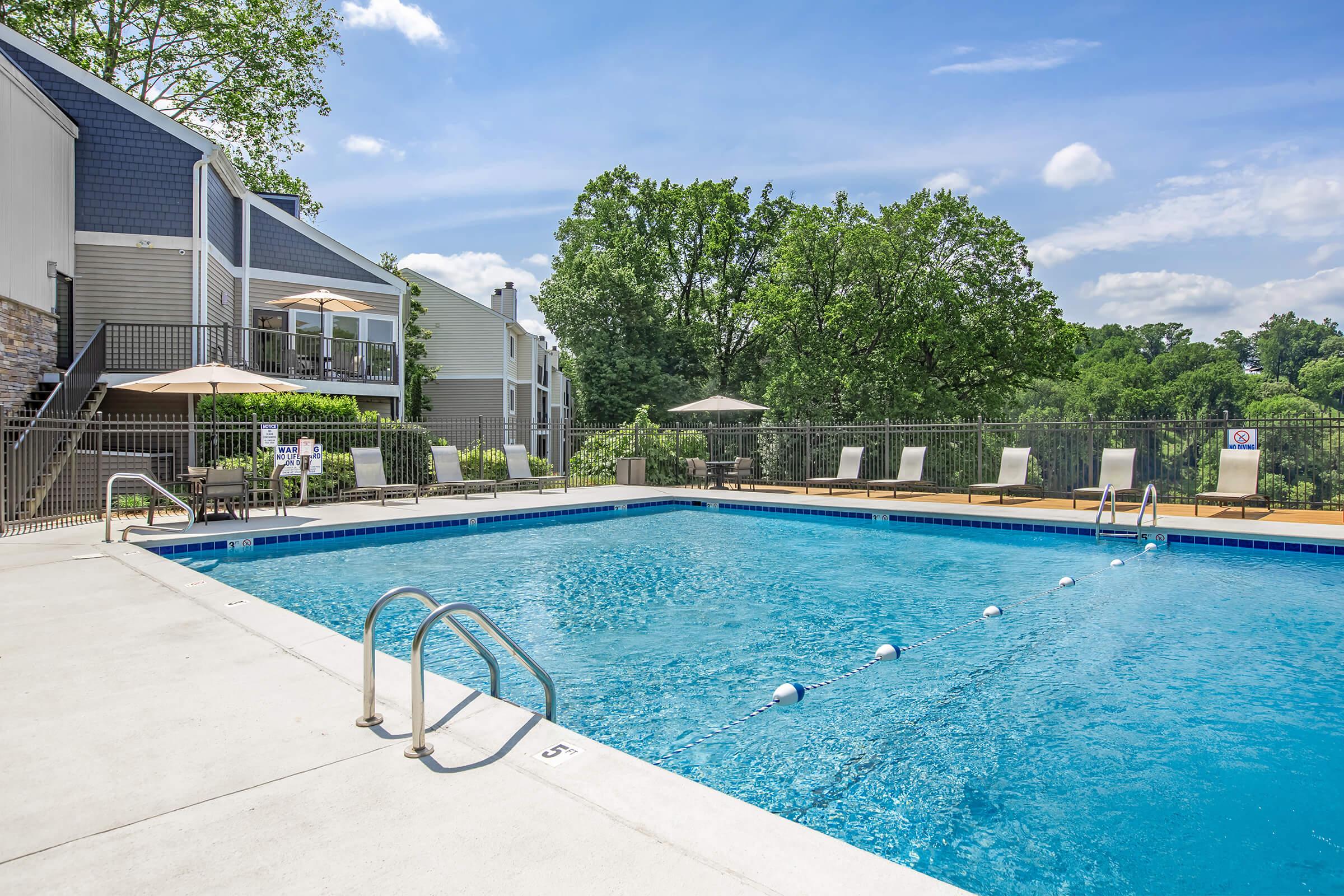 A sparkling blue swimming pool surrounded by lounge chairs and shaded umbrellas, set against a backdrop of green trees and a clear blue sky. Modern apartment buildings are visible in the background, creating a relaxing outdoor space suitable for leisure and recreation.