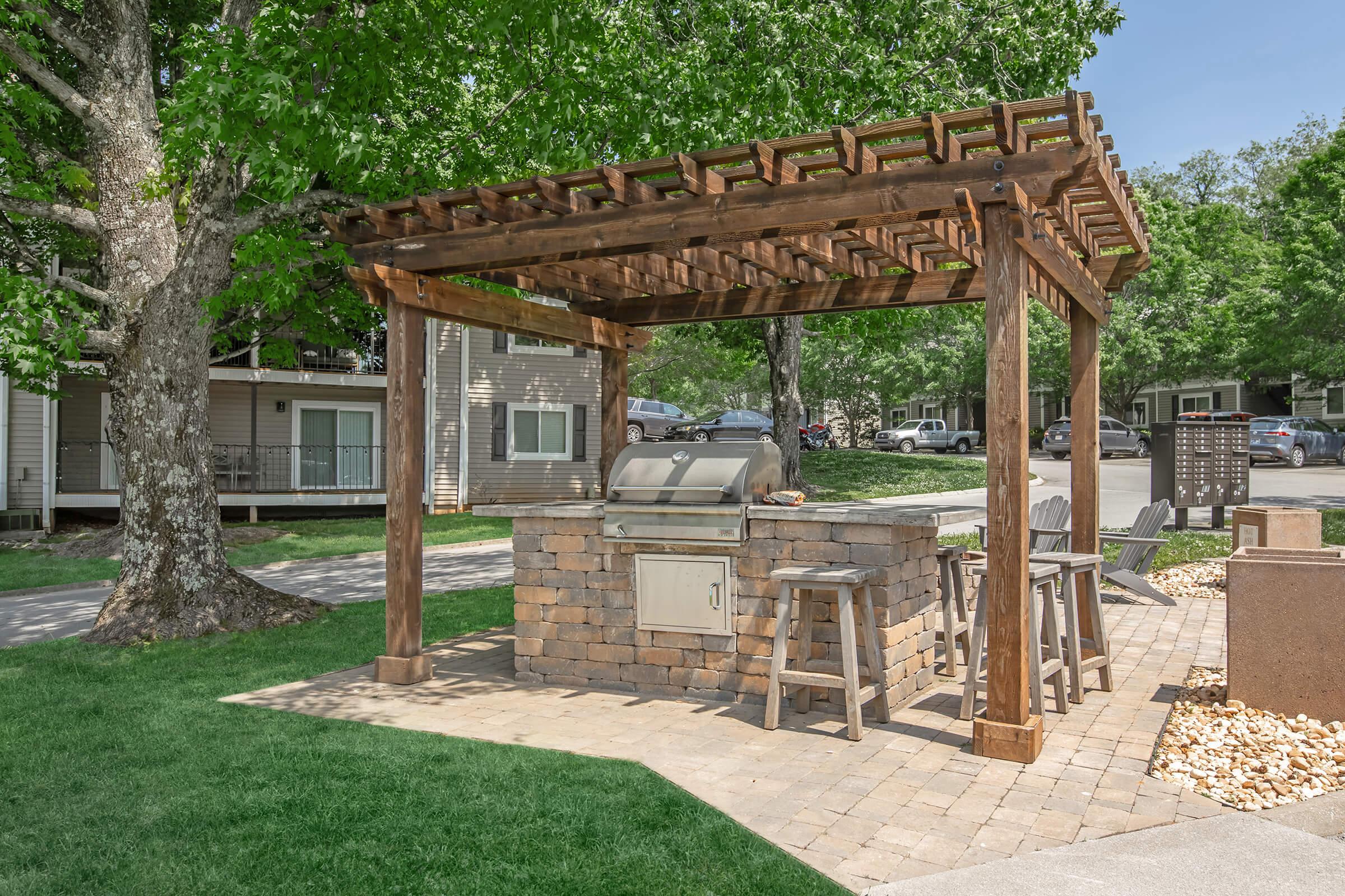 An outdoor grilling area featuring a stone grill under a wooden pergola. Surrounding the grill are three wooden barstools. The space is landscaped with grass and trees, and nearby are residential buildings and parked cars. Ideal for social gatherings and barbecues.