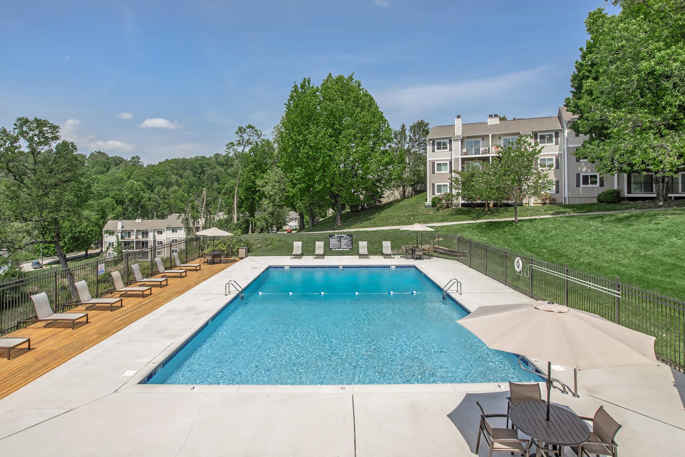A swimming pool surrounded by lounge chairs and umbrellas, set against a backdrop of green trees and residential buildings. The area is well-lit by a clear blue sky, creating a tranquil and inviting atmosphere for relaxation and recreation.