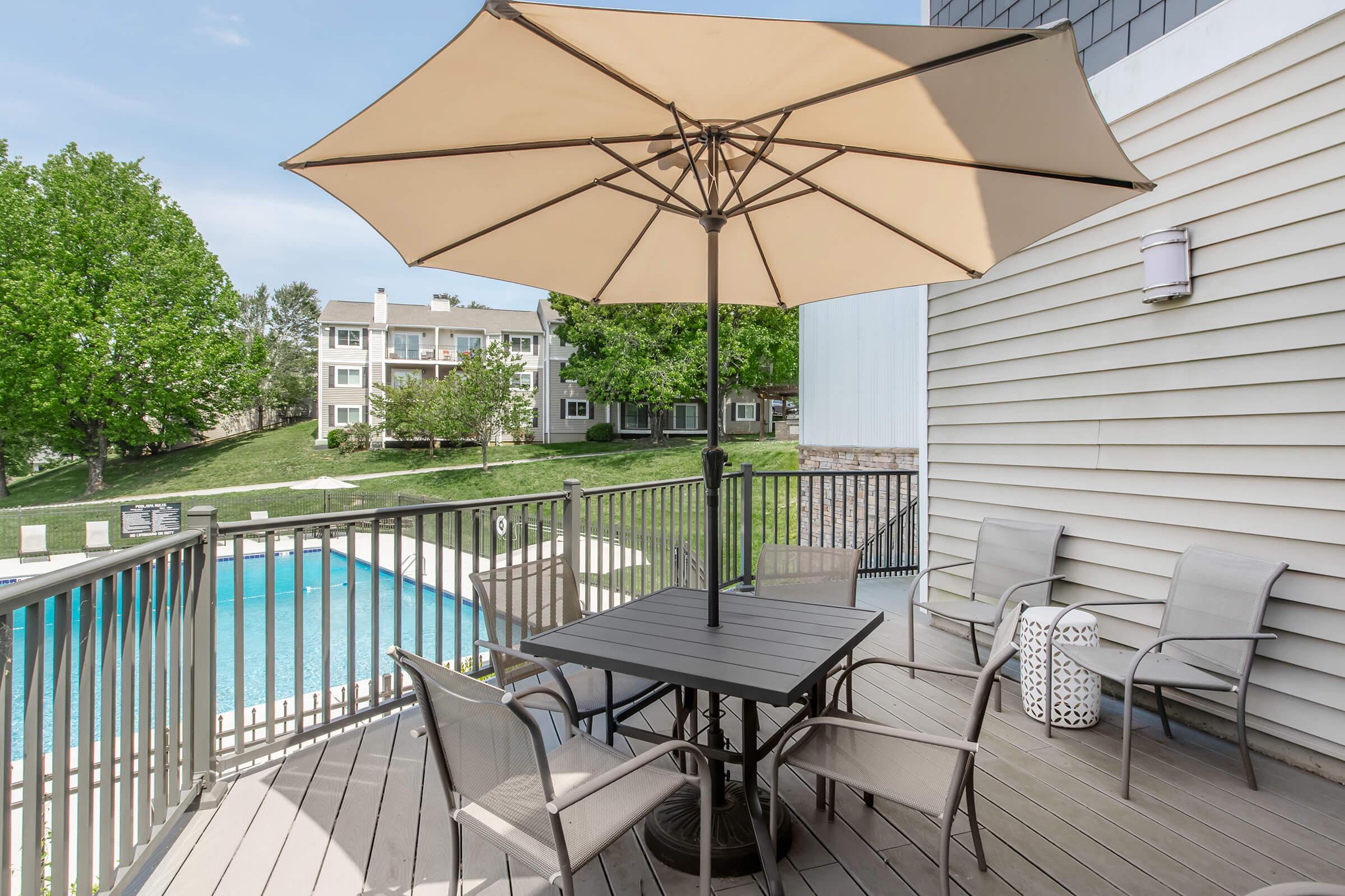 A patio area with a table and chairs under a large umbrella, overlooking a pool surrounded by green lawn and trees in a residential setting.
