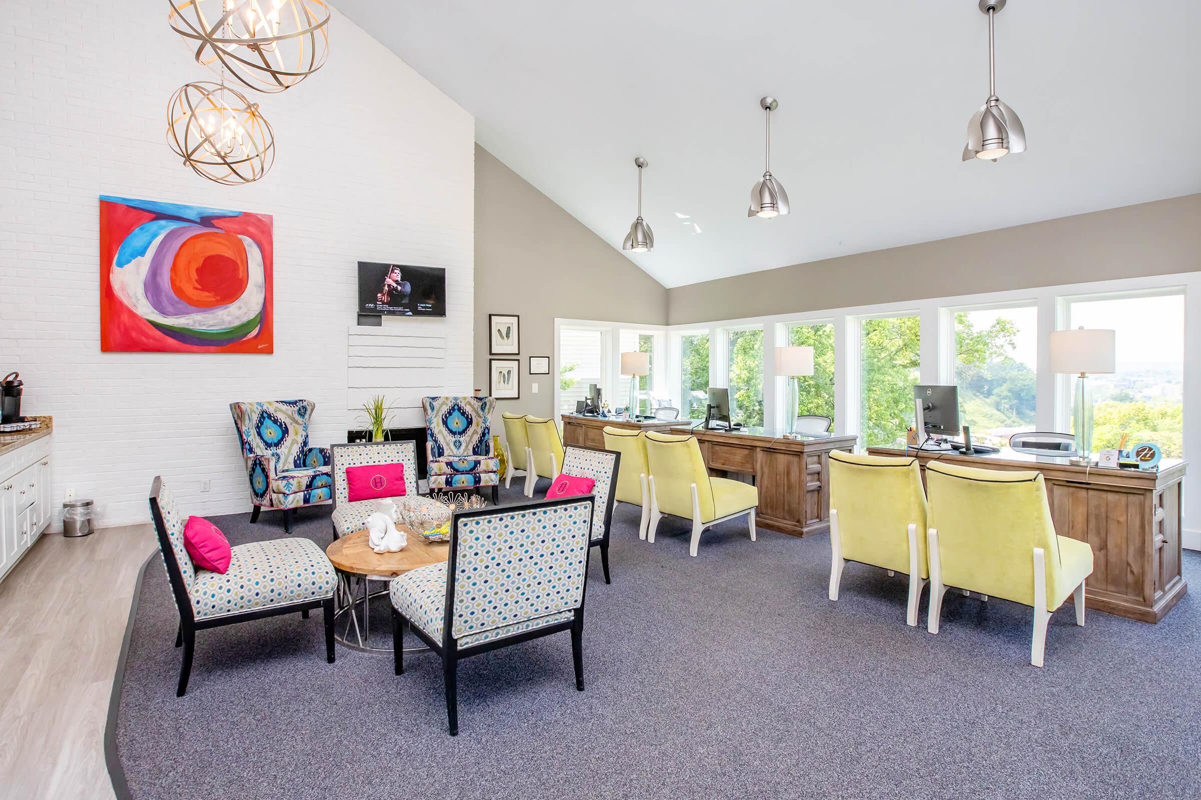 A modern lounge area featuring stylish furniture, including colorful chairs and a coffee table. Large windows provide a view of the outdoors, and there are workstations with computers. Decorative light fixtures hang from the ceiling, and a colorful piece of art is displayed on the wall. The space is bright and inviting.