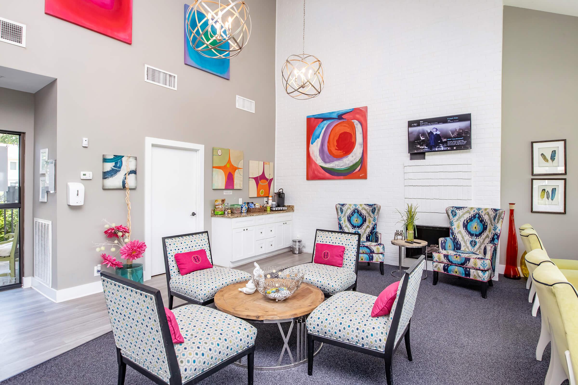 A modern living area featuring colorful abstract art on the walls, a circular wooden coffee table surrounded by four black chairs with pink cushions, and two patterned armchairs. A console table with decorative items sits against the wall, and a television is mounted above a white brick fireplace. Bright, inviting decor throughout.
