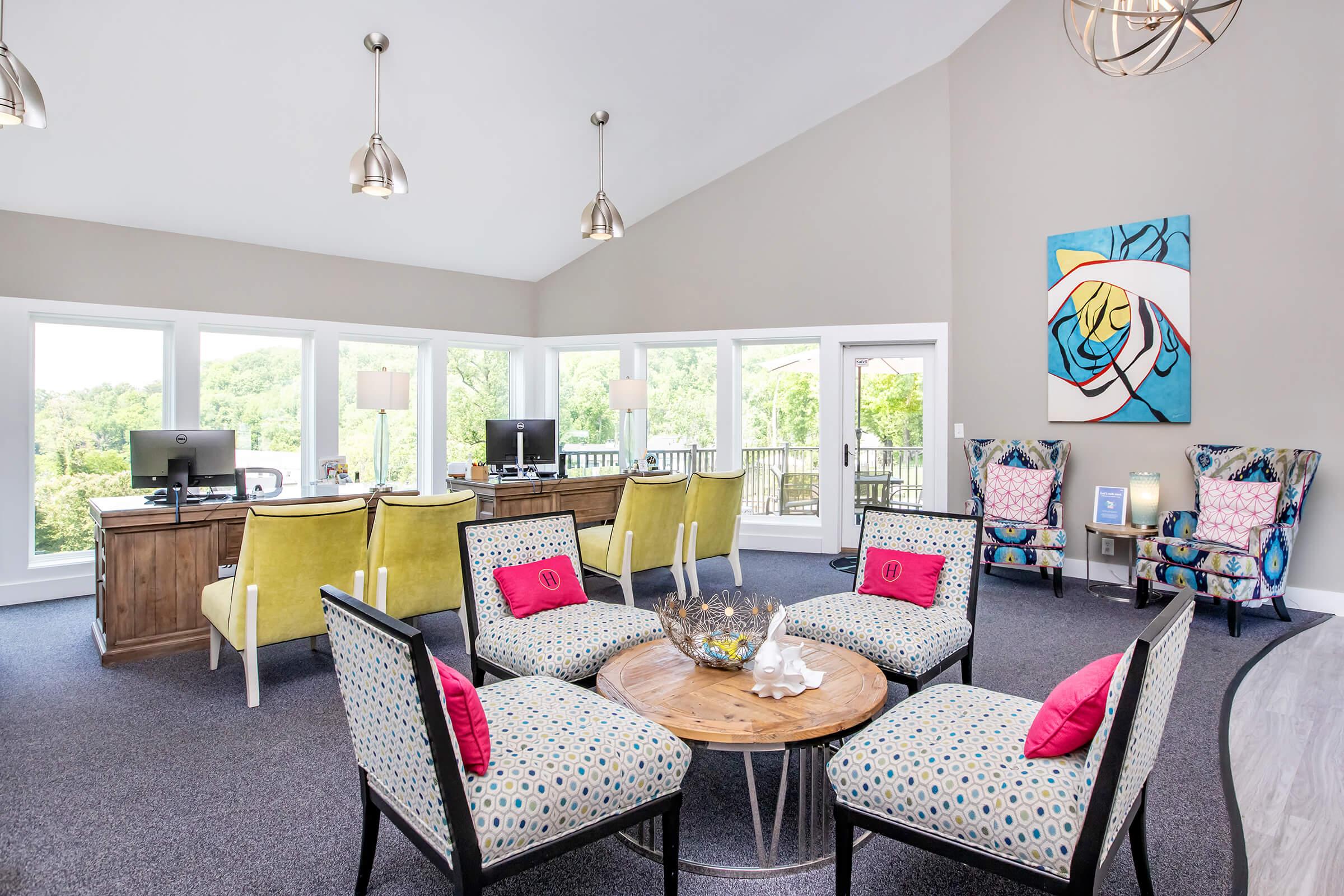A modern lounge area featuring stylish furniture, including colorful chairs and patterned seating around a wooden coffee table. Large windows offer natural light and views of greenery outside. Two computers are set up on a desk in the background, and decorative artwork hangs on the wall.