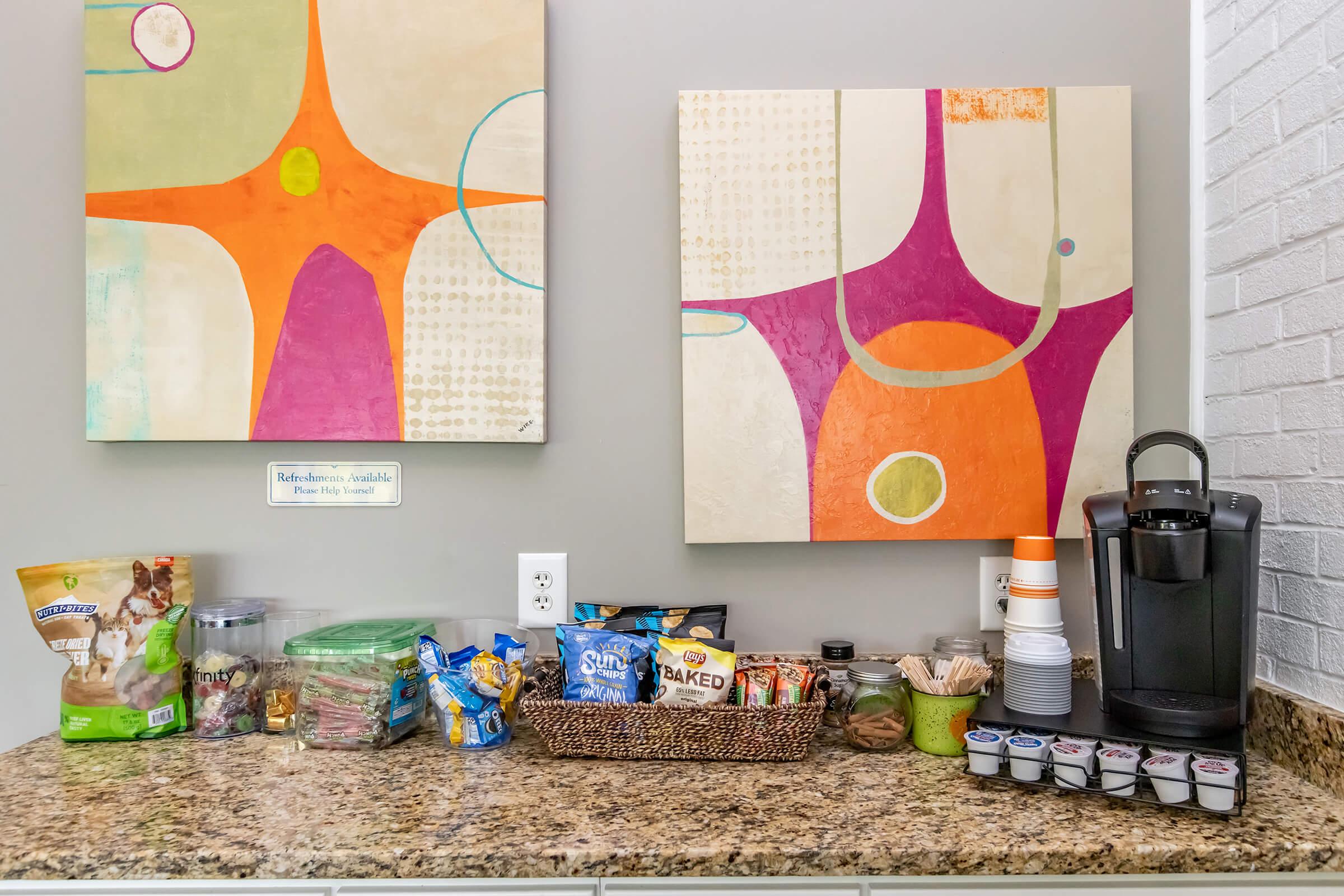 A snack station featuring a variety of packaged snacks, including chips, granola bars, and nuts, arranged on a granite countertop. Nearby, an assortment of coffee pods and cups is displayed alongside two colorful abstract artworks on the wall.