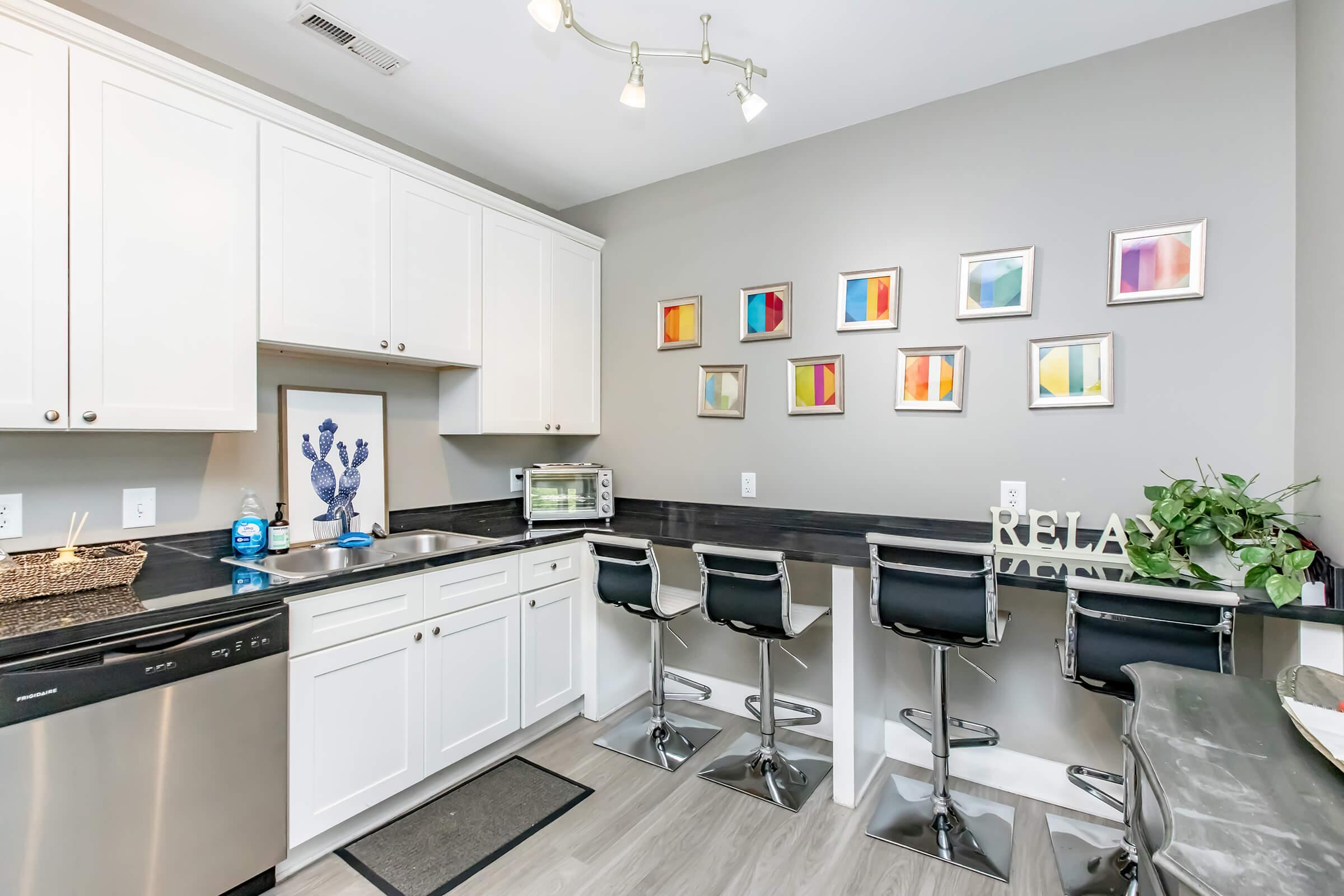 Modern kitchen featuring white cabinetry, a black countertop, stainless steel appliances, and a decorative back wall. There are four bar stools at a breakfast counter, and colorful framed artwork hangs above. A small plant is on the counter, adding a touch of greenery to the space.
