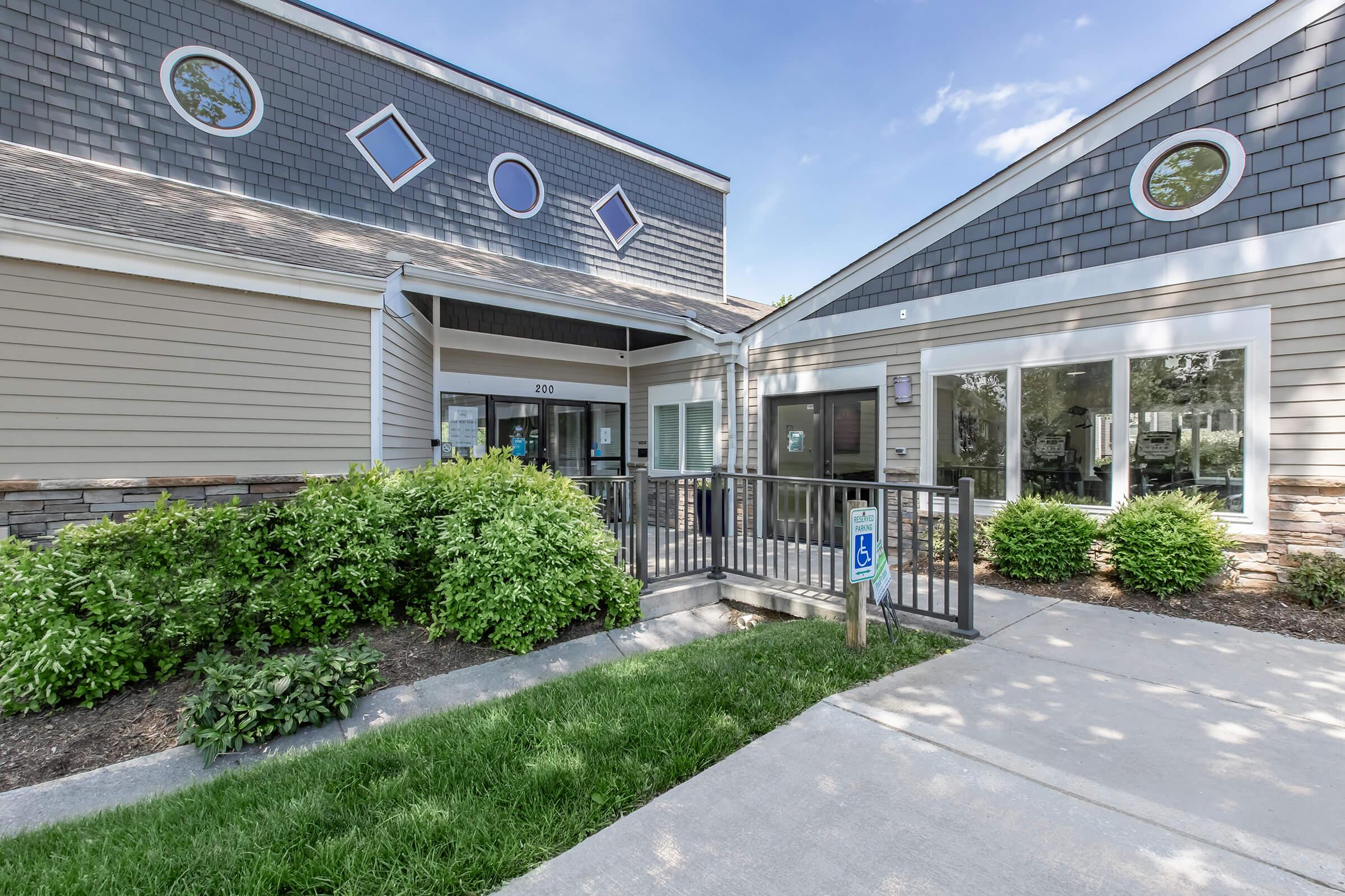Exterior view of a modern, single-story building with a textured facade, featuring large windows and decorative elements. A pathway with a ramp leads to the entrance, surrounded by landscaped greenery and small bushes. Clear blue sky overhead.