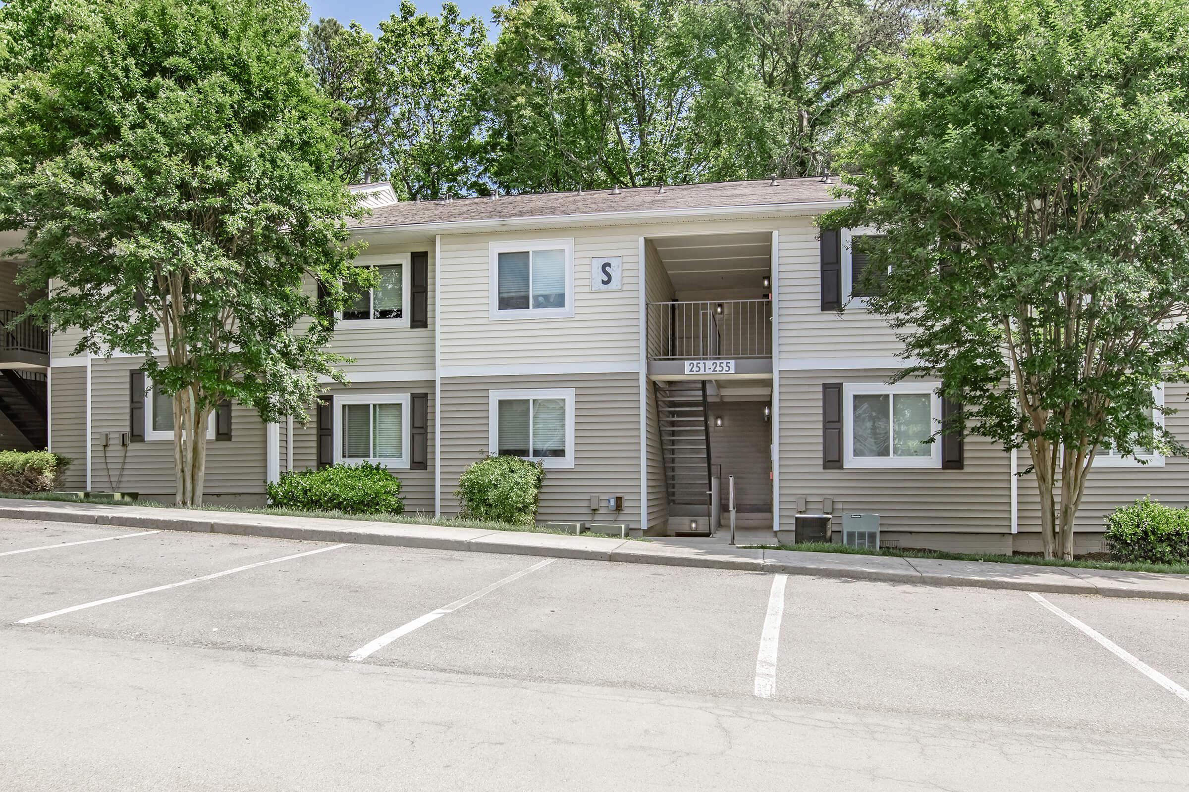 Two-story apartment building with light-colored siding, surrounded by trees and shrubs. Parking spaces are positioned in front, and the entrance features a staircase. Windows are neatly aligned on both floors, showcasing a well-maintained residential area.
