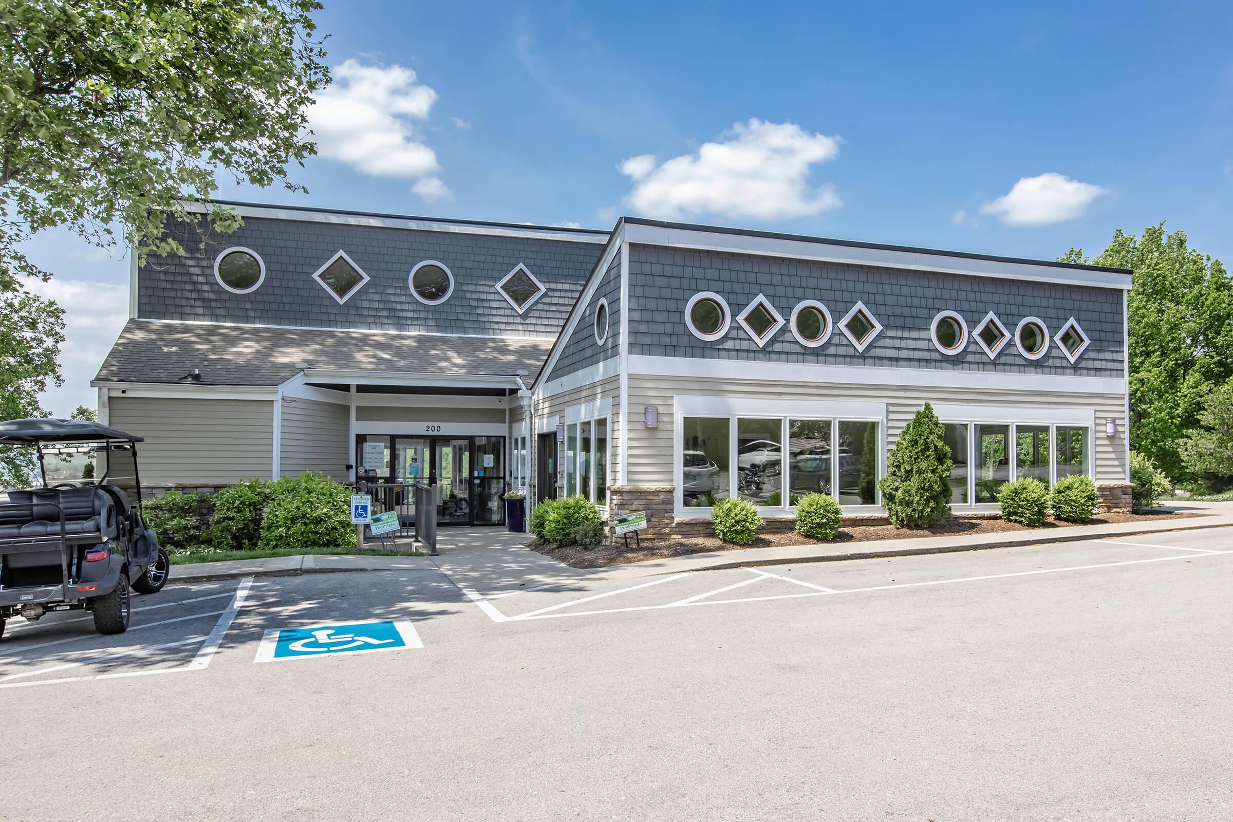 A modern building with gray siding and decorative diamond-shaped windows. It features a landscaped entrance, parked vehicle, and a designated accessible parking space in front. The sky is clear with a few clouds, showcasing a bright and inviting atmosphere.