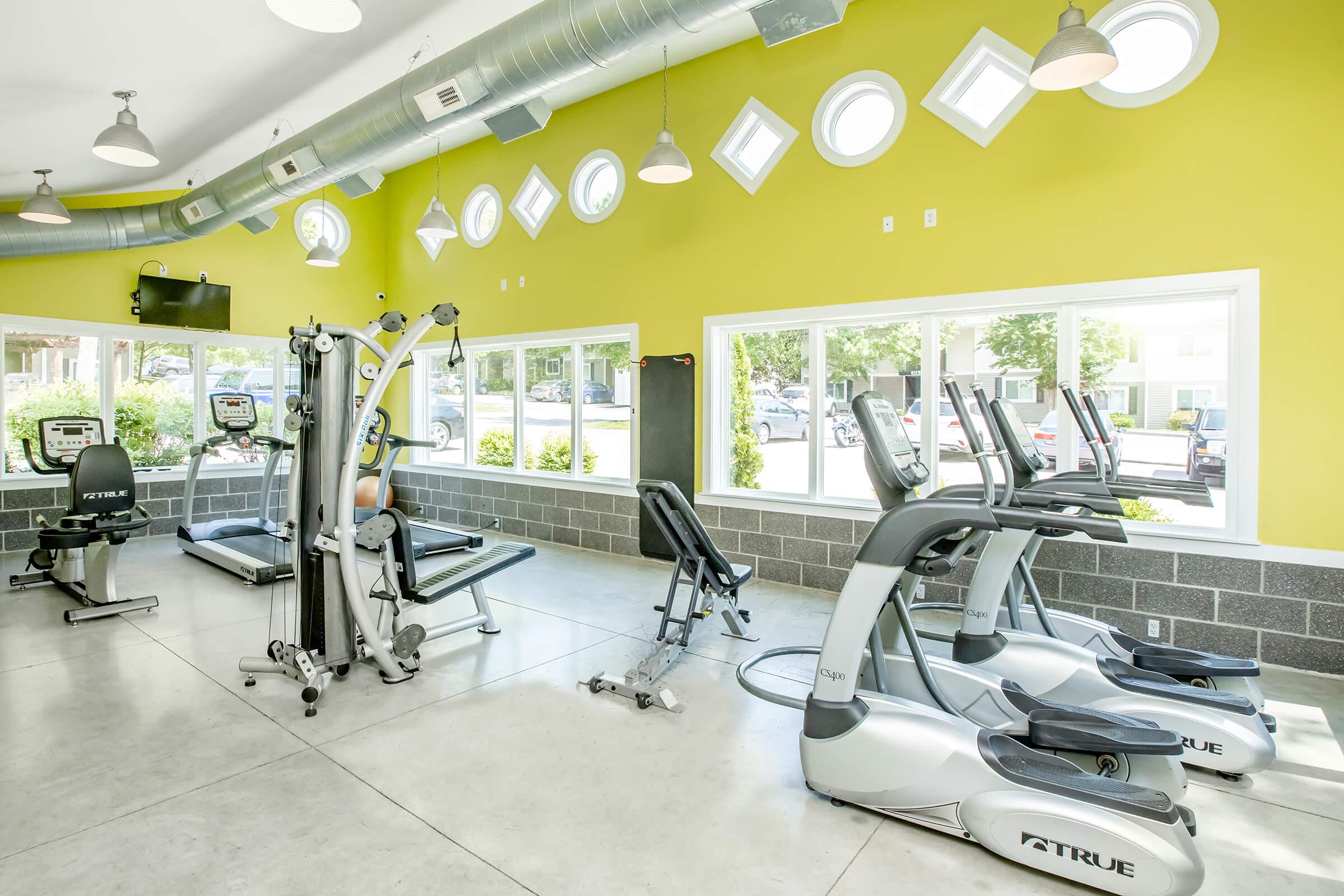 A bright and modern fitness room featuring various exercise equipment, including ellipticals, stationary bikes, and strength-training machines. The walls are painted a vibrant yellow, and large windows allow natural light to fill the space, creating an inviting atmosphere for workouts.