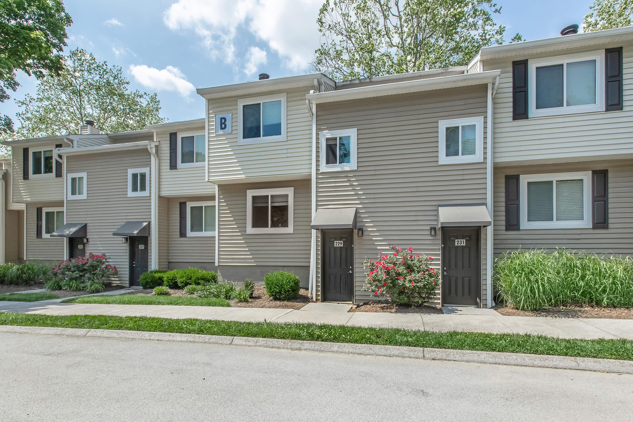 A row of modern townhouses with light gray siding, each featuring a front door and small window. The units have black shutters and awnings, with flowering bushes and green shrubs in front. The scene is set on a sunny day, with a clear blue sky and some clouds visible.