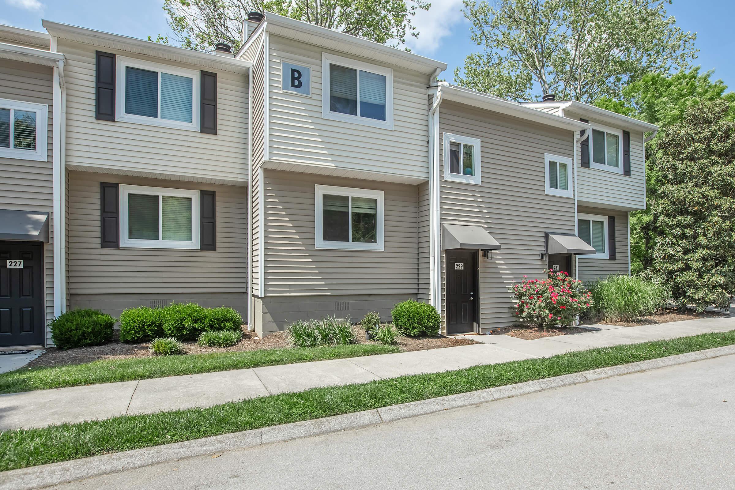 A row of three residential townhouses with beige siding and black doors. Each unit has windows with white trim and small awnings. The front yards are landscaped with shrubs and flowers, and a paved sidewalk runs in front of the houses, bordered by a green lawn. Bright blue sky visible above.