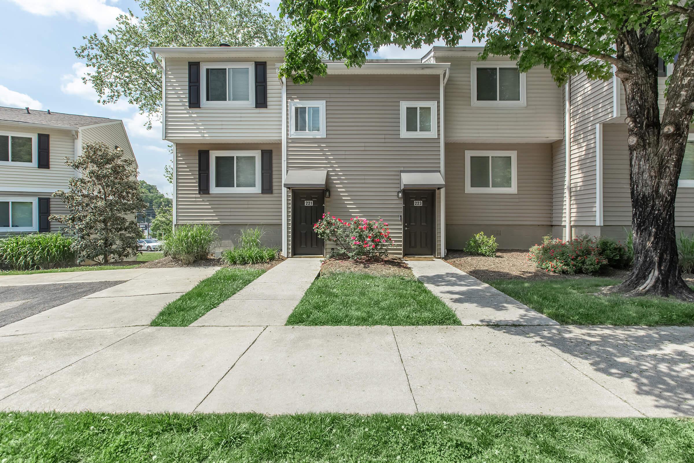 A two-story residential building featuring two front doors, surrounded by green grass and flower beds. There are neatly trimmed shrubs and trees nearby, under a clear sky with soft clouds. The pathway leads to the entrance of the units, showcasing a well-maintained exterior.