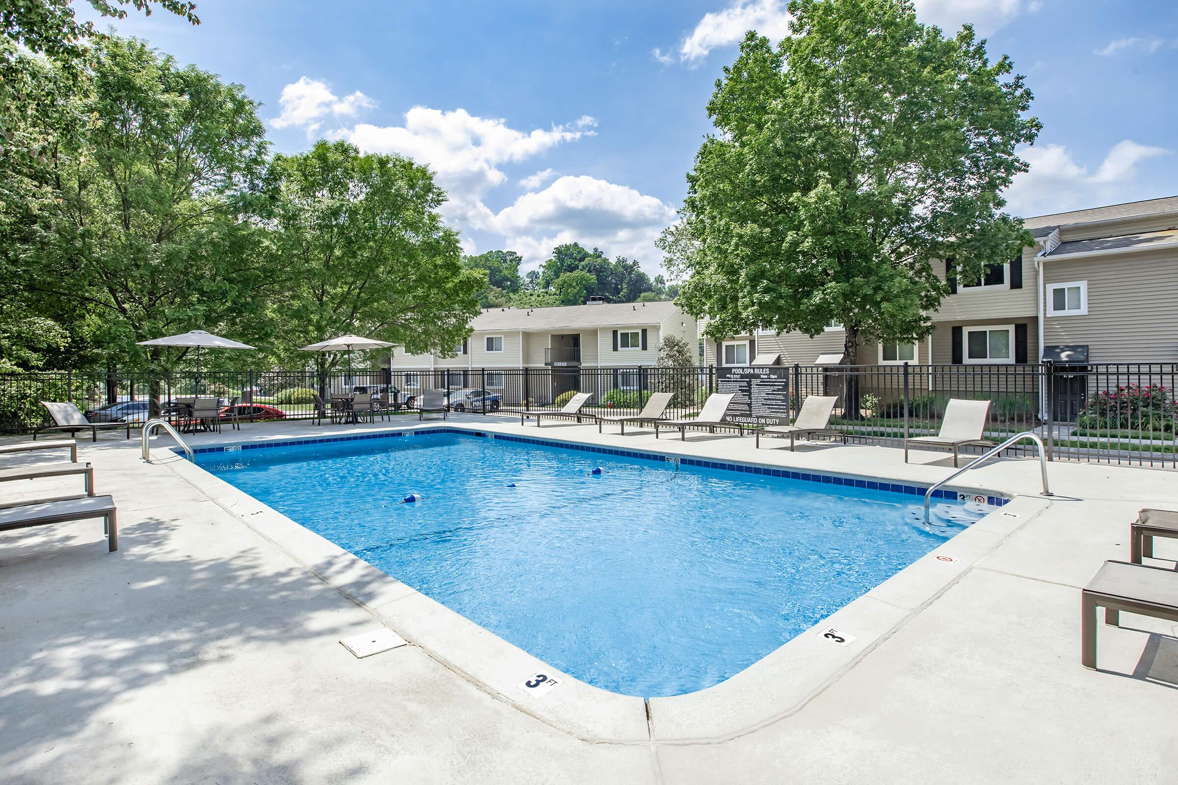 Swimming pool area with blue water surrounded by lounge chairs and umbrellas. Lush green trees provide shade, and several apartment buildings are visible in the background under a partly cloudy sky. The pool is marked with depth indicators along the edges.