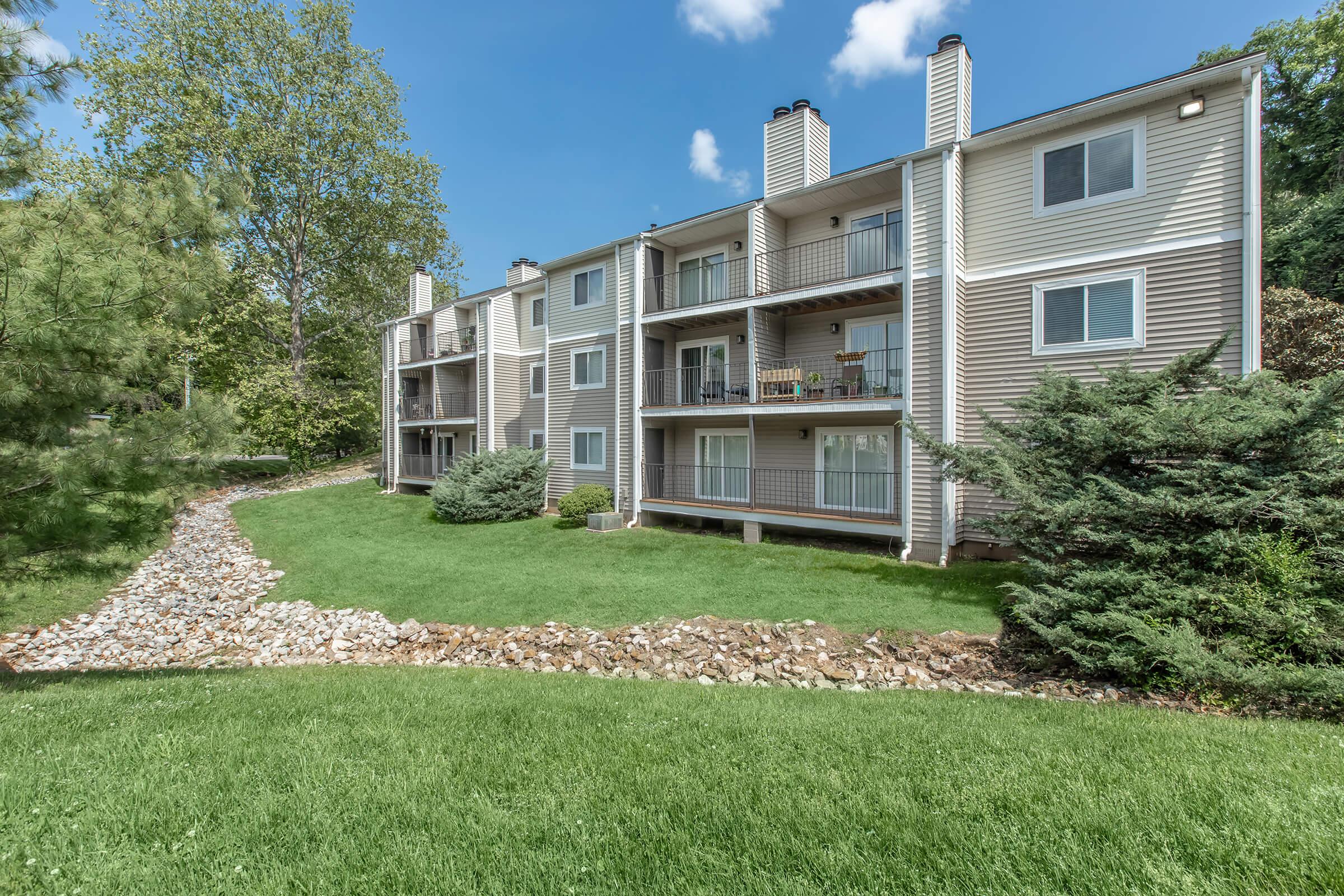 A multi-unit apartment building with several balconies, situated in a lush green landscape. The ground features a well-maintained lawn and a pathway made of stones, while trees provide shade around the property under a blue sky with scattered clouds.