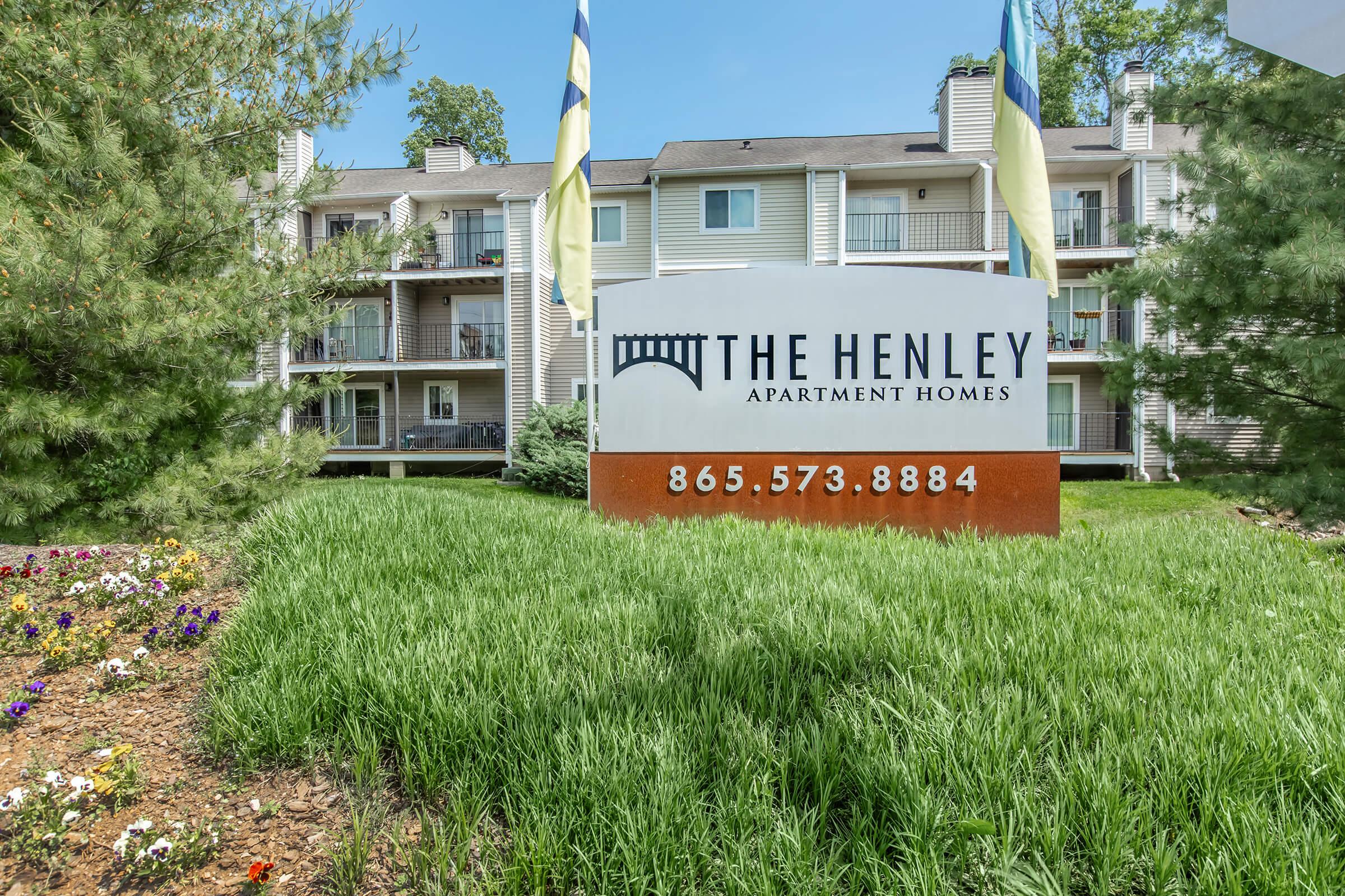 Sign displaying "The Henley Apartment Homes" with contact number 865-573-8884, surrounded by green grass and flower beds, in front of a multi-story apartment building with balconies.