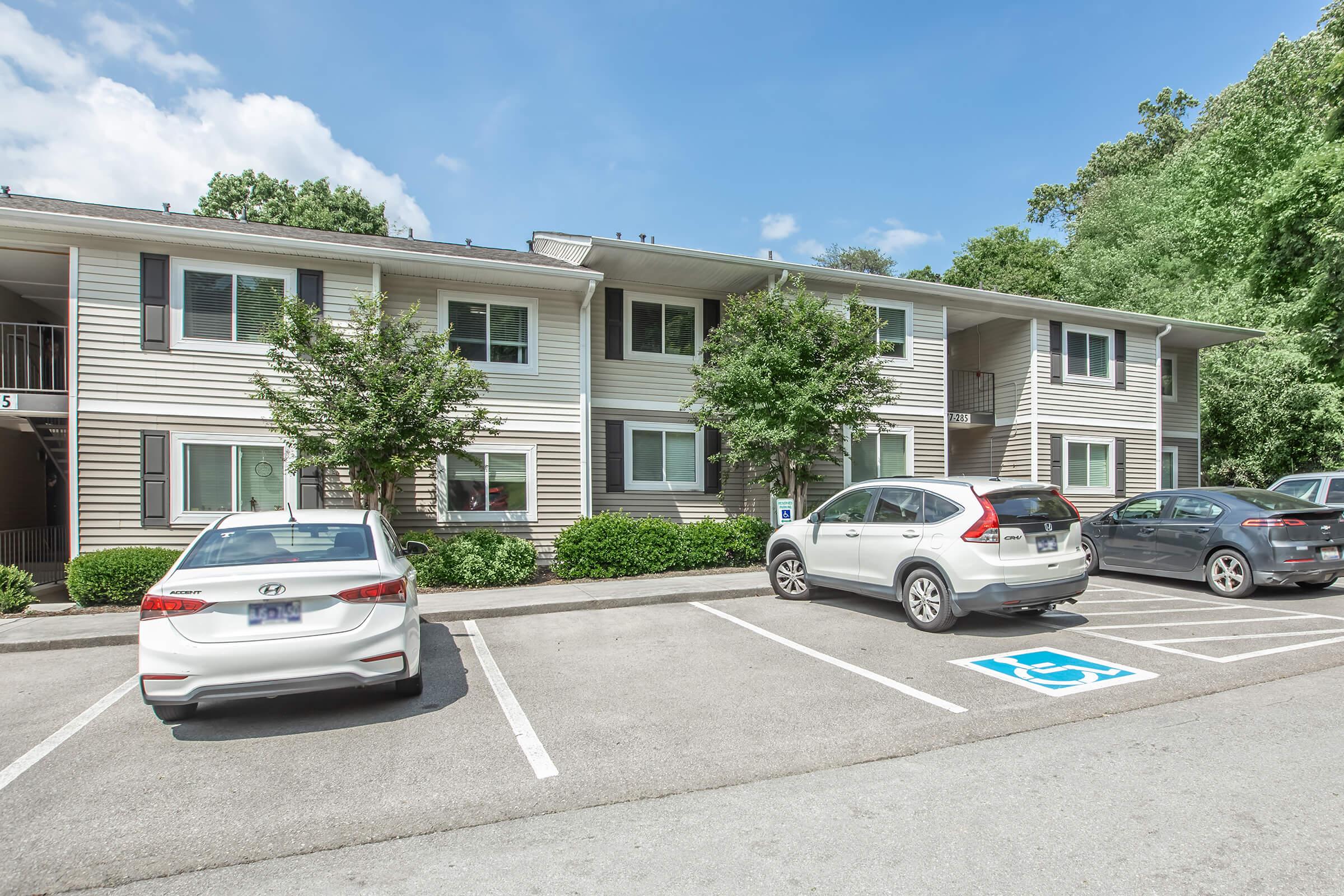 A two-story apartment building with light-colored siding surrounded by trees. There are several parked cars in the foreground, including a white sedan and an SUV. A designated handicapped parking space is visible. The sky is partly cloudy, indicating a bright day.