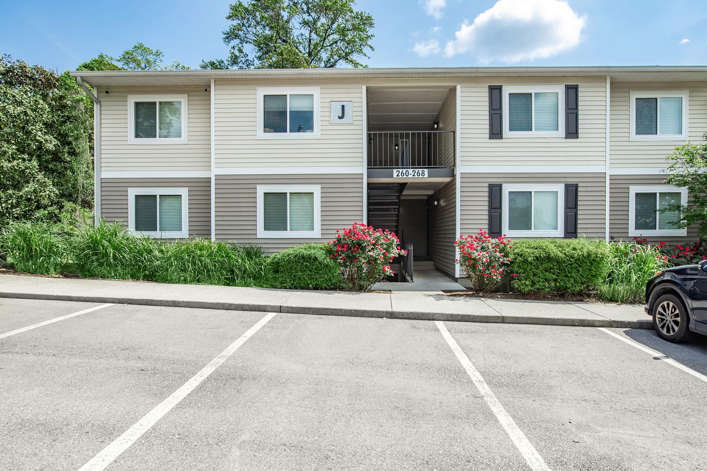 Exterior view of a residential apartment building with two stories, featuring beige and gray siding. The entrance is marked with the numbers 264-268, surrounded by green bushes and blooming flowers. A parking lot is visible in the foreground, with a black car parked nearby. Trees and blue sky in the background.