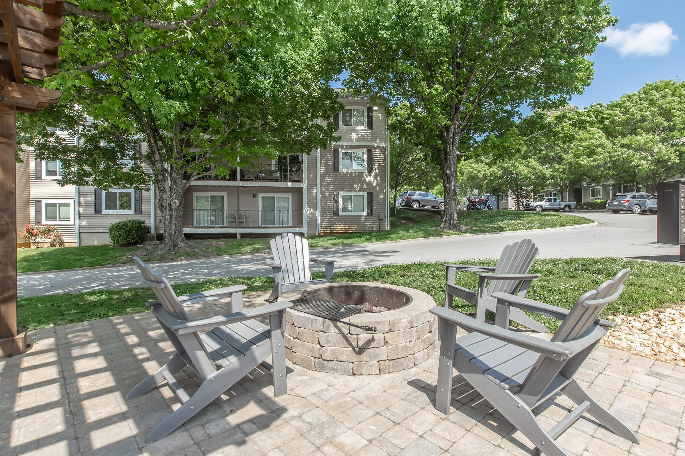 A cozy outdoor space featuring a circular stone fire pit surrounded by four gray Adirondack chairs. Lush green trees provide shade, and an apartment building is visible in the background along with a paved pathway and parked cars. The scene is bright and inviting, perfect for gatherings.