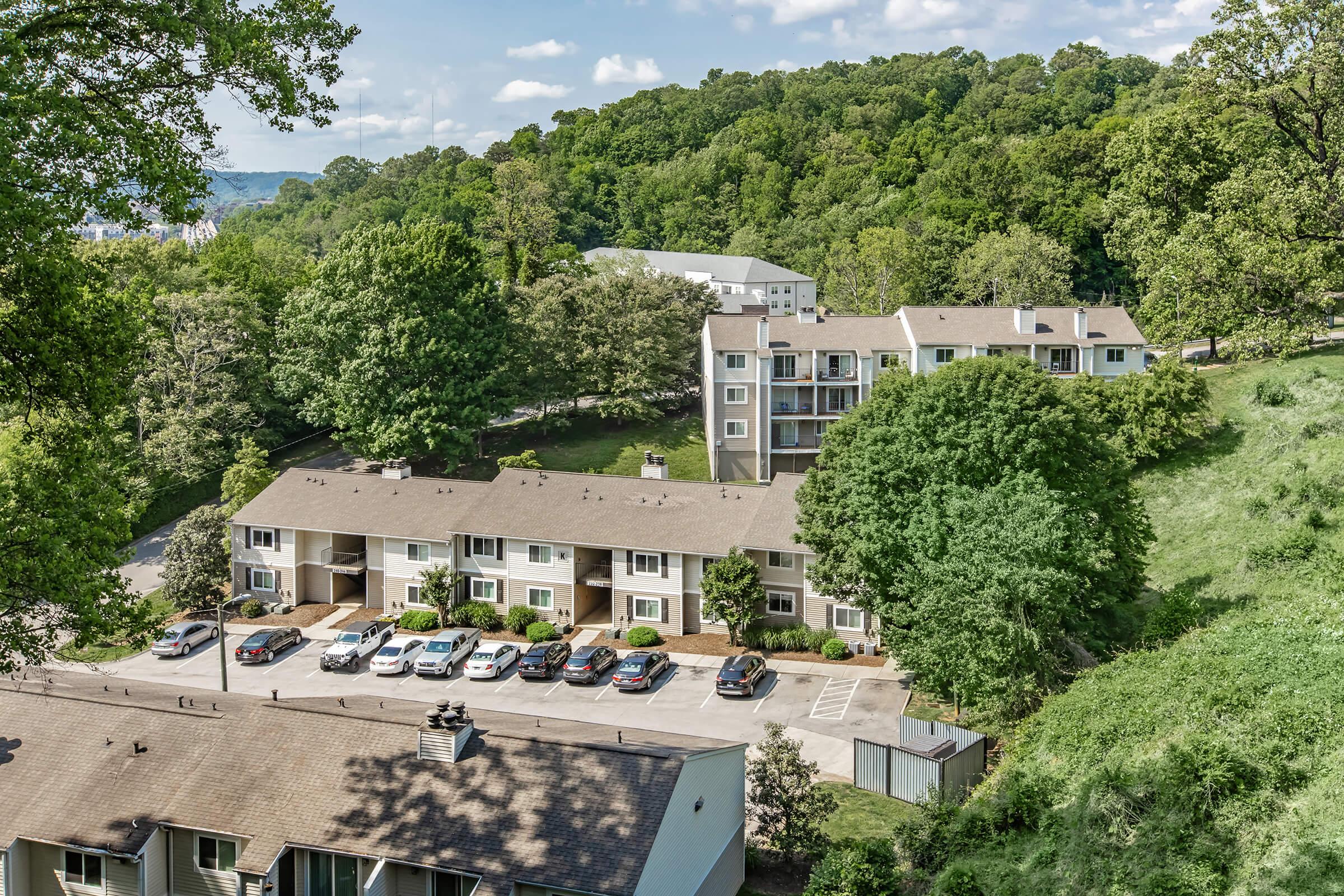 Aerial view of a residential area featuring several apartment buildings surrounded by lush greenery. The foreground includes a lower building with parking spaces filled with cars, while a larger building is visible in the background, nestled among trees on a hillside. Blue skies with a few clouds above.