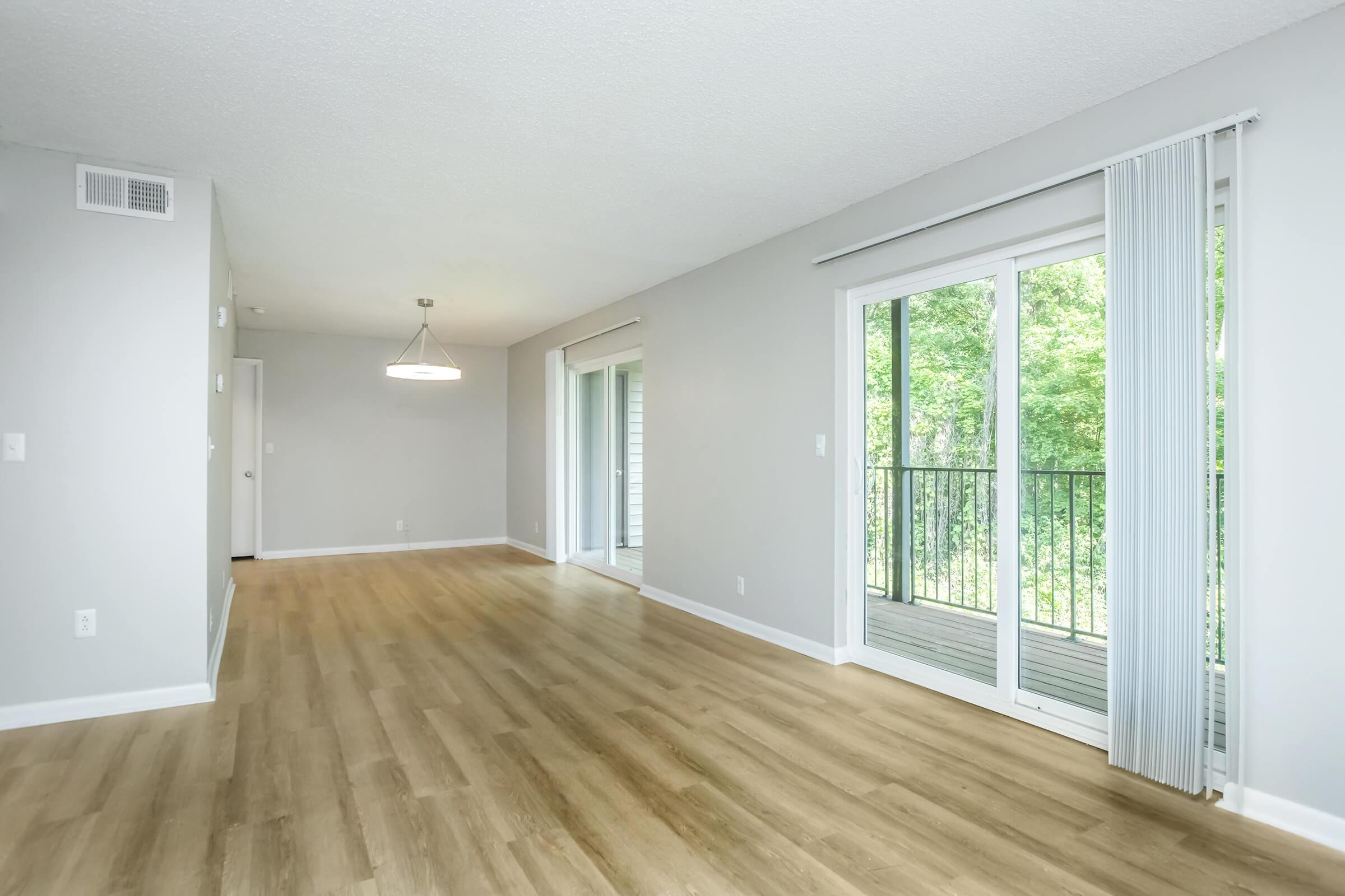 A spacious living room with light-colored walls and hardwood flooring. Large sliding glass doors open to a balcony overlooking green trees, allowing natural light to fill the space. A modern light fixture hangs from the ceiling, adding a contemporary touch to the inviting atmosphere.