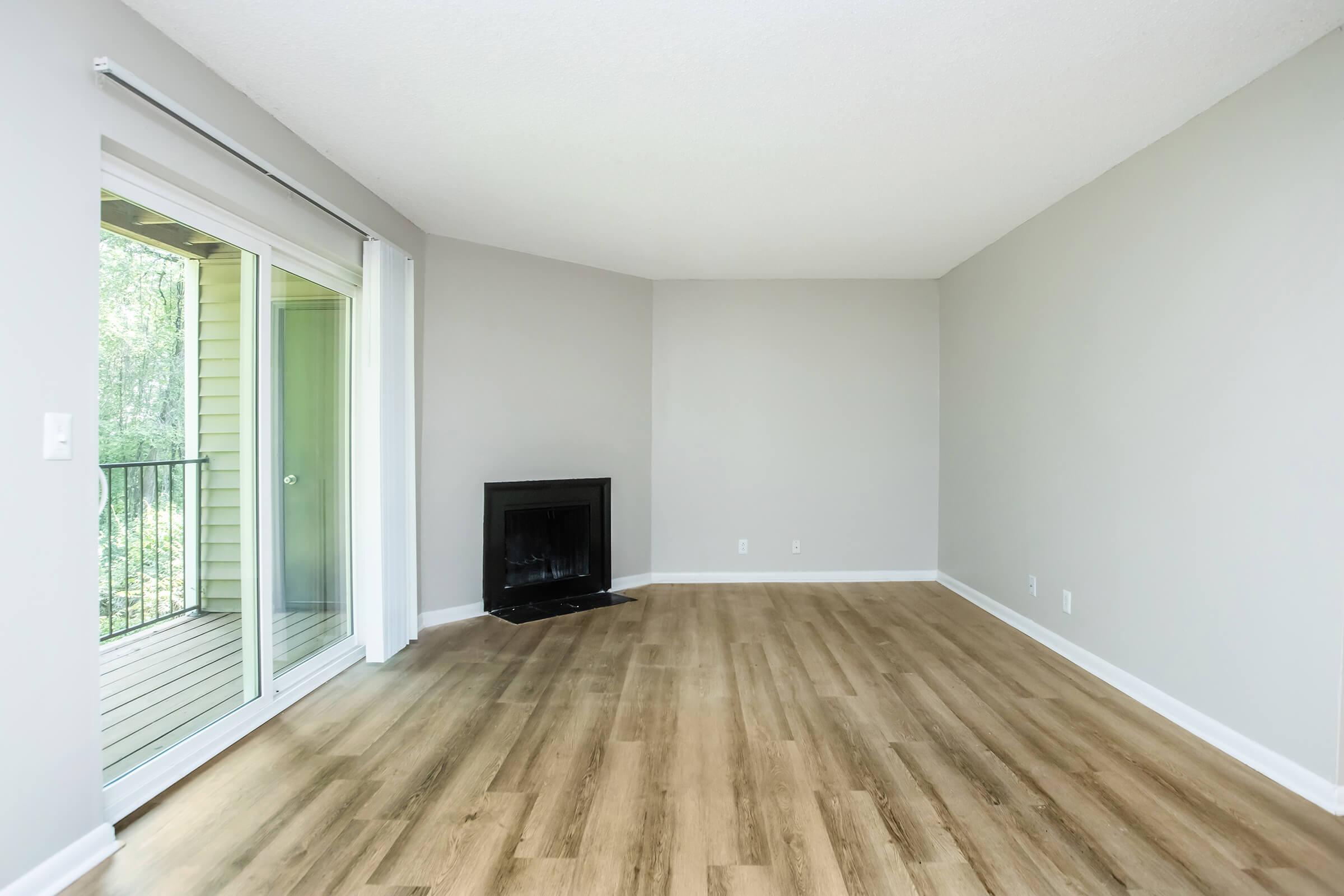 Empty room with light gray walls and wooden flooring. A black fireplace is positioned on one side, and large sliding glass doors lead to a balcony with a view of greenery outside. The space has a simple, modern design with no furniture present.