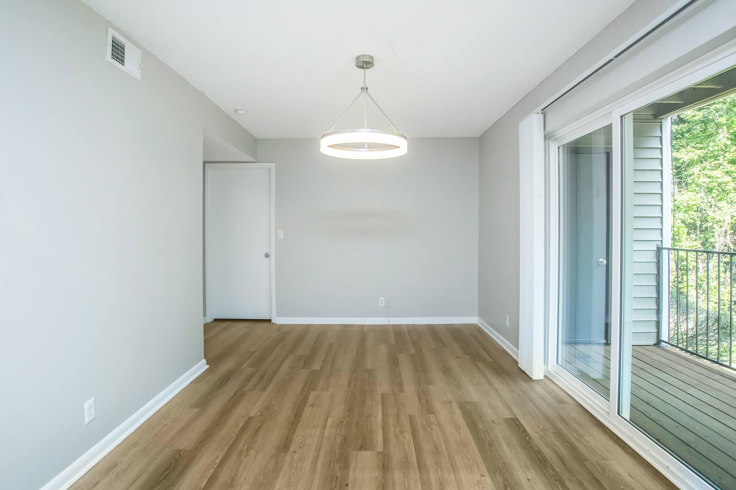 A spacious, modern room with light gray walls and new wood laminate flooring. A contemporary pendant light hangs from the ceiling, providing soft illumination. There are sliding glass doors leading to a small balcony surrounded by greenery, enhancing the natural light and outdoor connection.