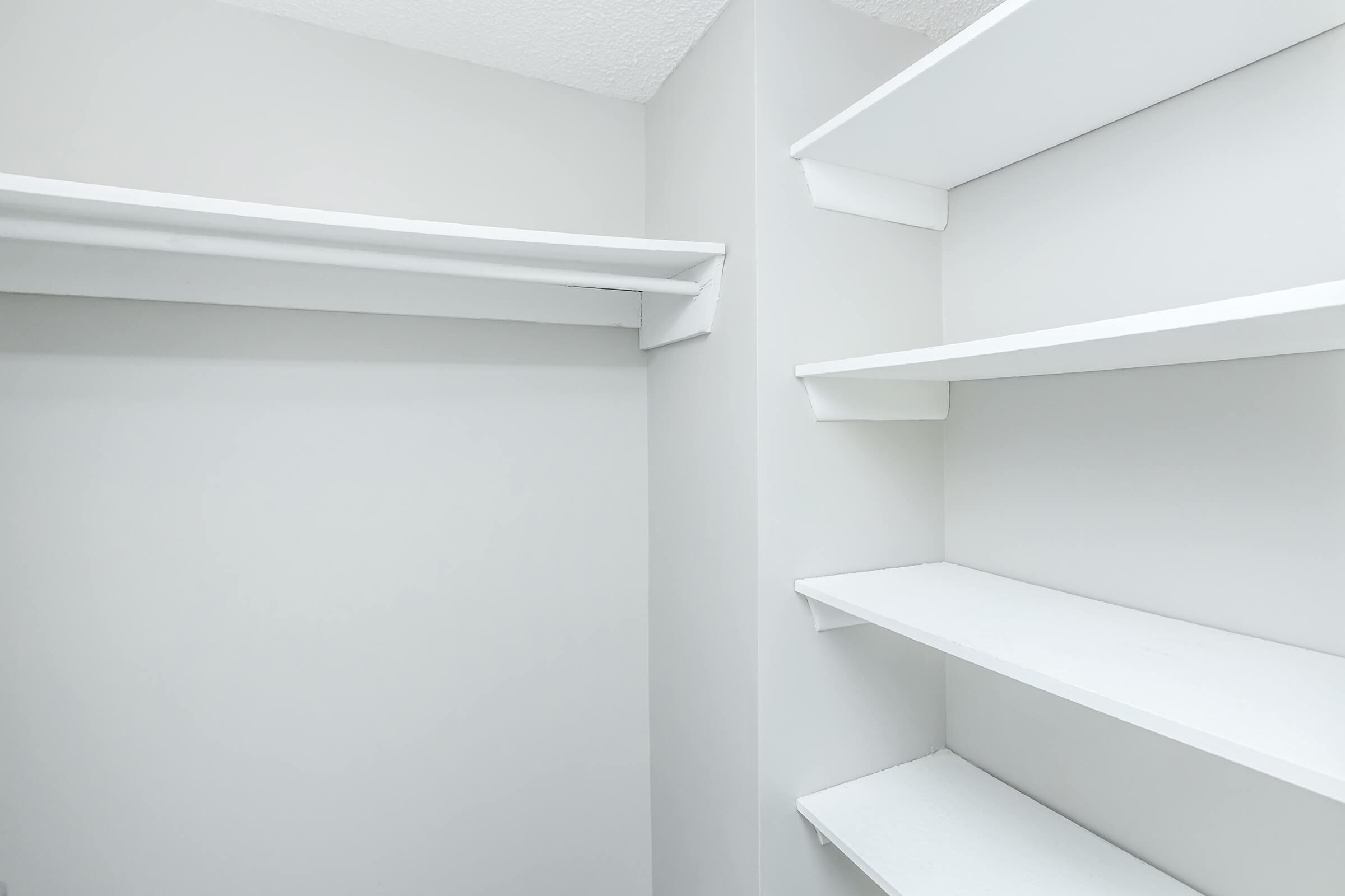 Empty closet with white shelves against a light gray wall. The left side features a hanging rod, while the right side has three open shelves. The overall aesthetic is clean and minimalist, creating a spacious appearance.