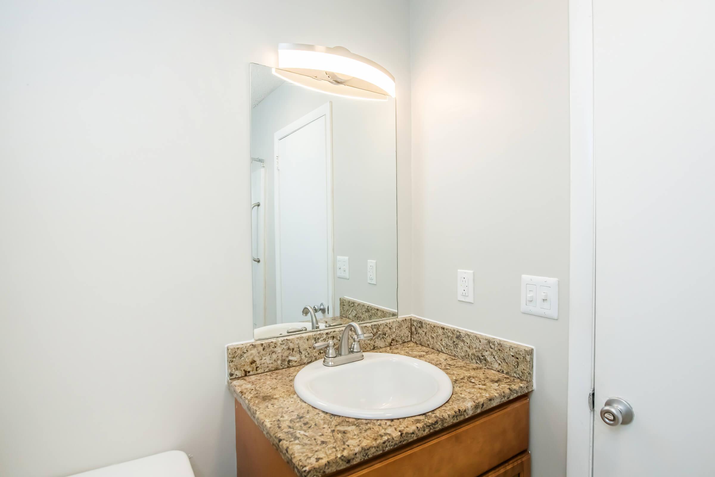 A modern bathroom with a light-colored granite countertop and a circular sink. The mirror above the sink is well-lit by a sleek, contemporary light fixture. There is a white towel rack and light gray walls, creating a clean and minimalist aesthetic. The door is partially visible on the right.