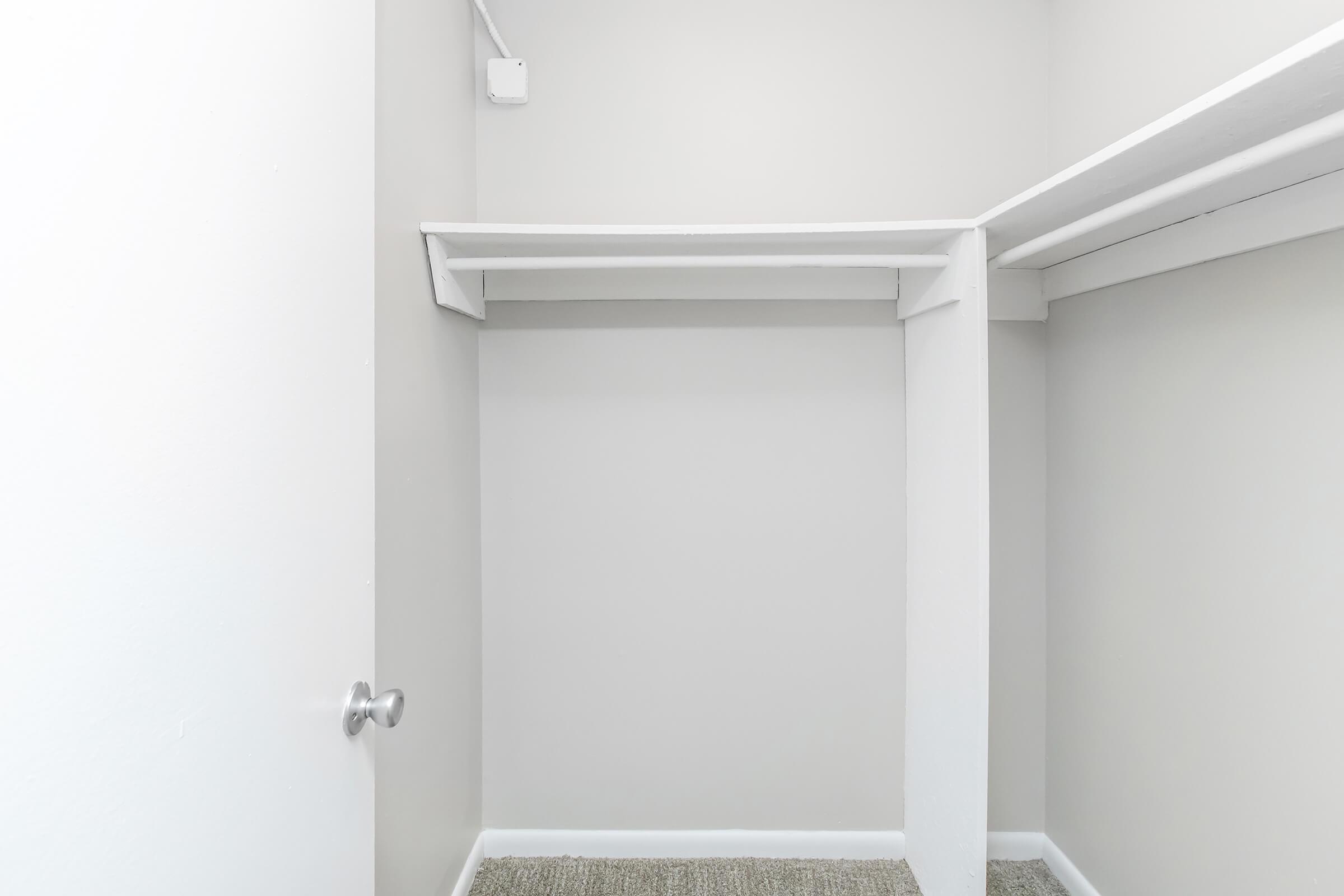 An empty closet with light gray walls and a light gray carpet. There are two shelves on the upper part of the wall, and a door slightly ajar on the left side. The closet is spacious and well-lit, providing a clean and organized storage space.