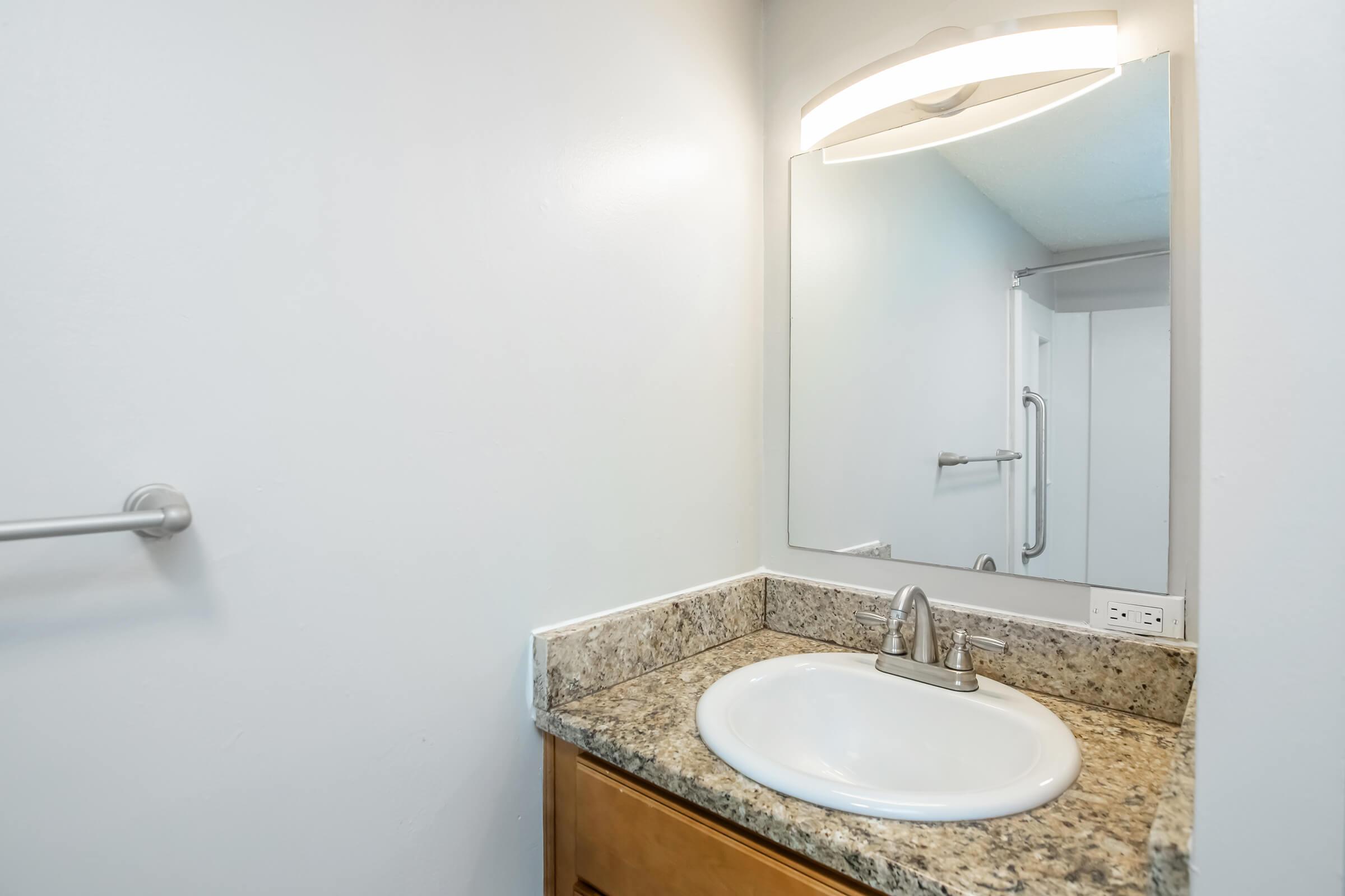 A modern bathroom sink area featuring a white basin, a polished faucet, and a granite countertop. Above the sink, there is a rectangular mirror with soft lighting. A towel bar is mounted on the left wall, and a shower area is partially visible in the background. The walls are painted in a light color.