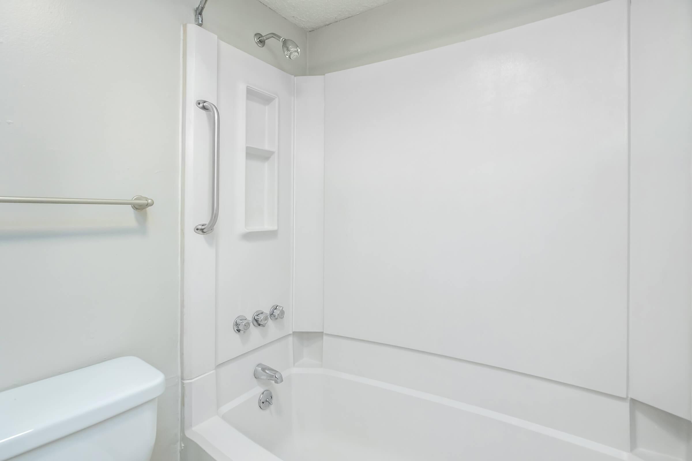 A clean and modern bathroom featuring a white bathtub and shower combo, with a grab bar for safety. The walls are light-colored, and there is a toilet nearby. A built-in shelf is visible in the shower area for toiletries.