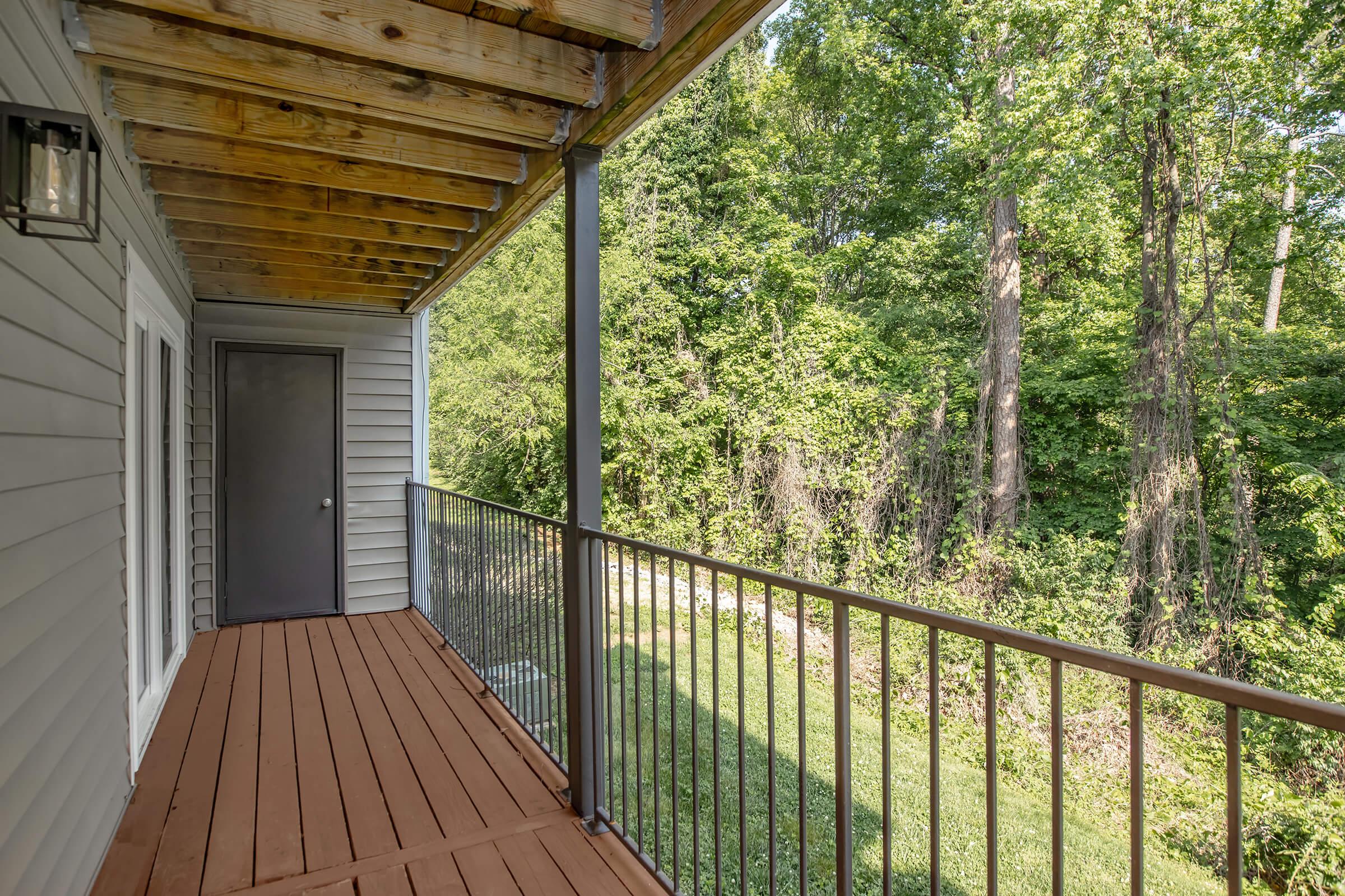 A wooden balcony with metal railing, featuring exposed beams overhead and a door leading inside. The balcony overlooks a lush green landscape with trees and grass, creating a serene natural setting.