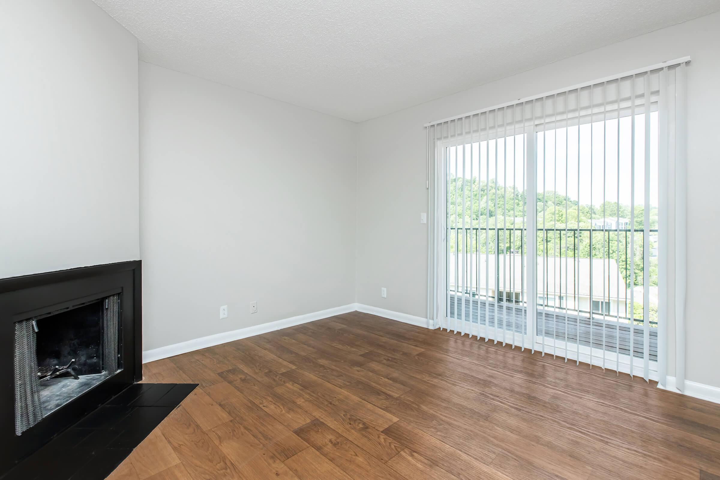 A spacious, modern living room with light gray walls, hardwood flooring, and a black fireplace. Large sliding glass doors with vertical blinds lead to a balcony, offering a view of greenery outside. The room is empty, creating a clean and inviting atmosphere.
