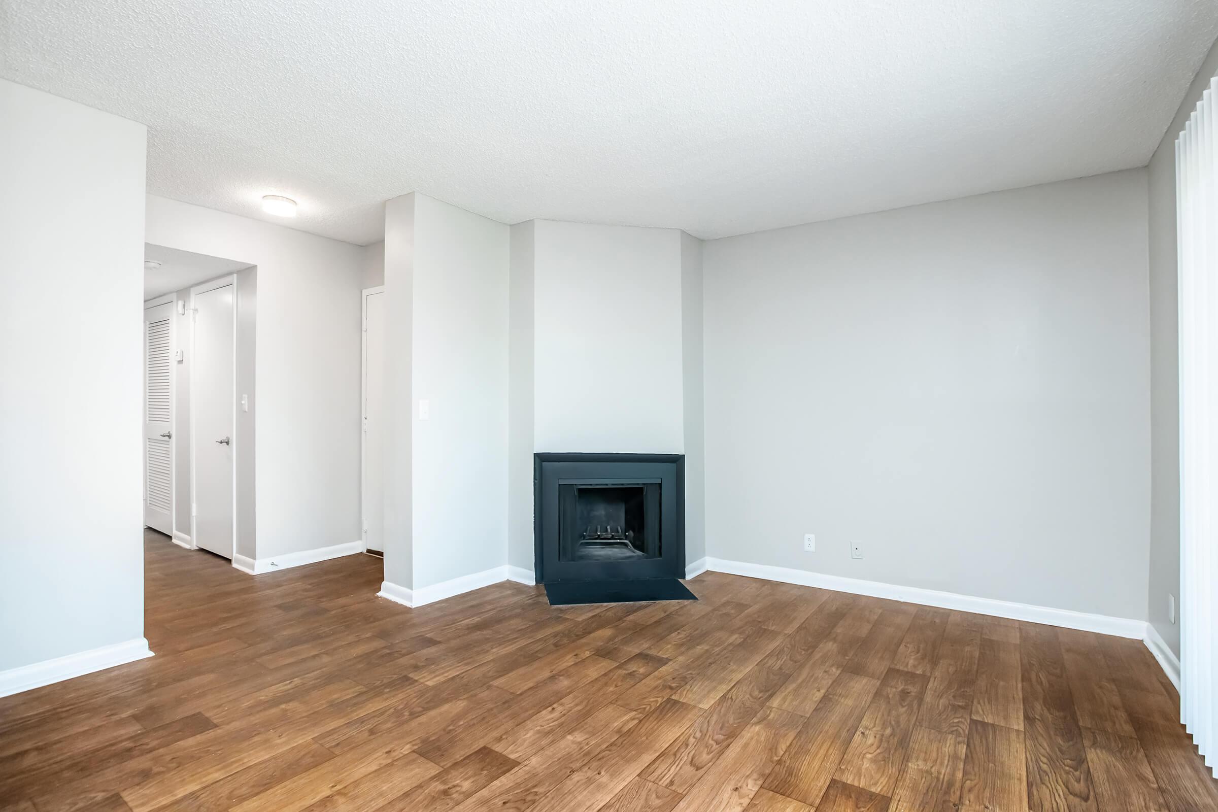 A spacious, empty living room featuring a dark fireplace on one wall, light gray walls, and hardwood flooring. The room is bright with natural light coming through a nearby window, and a doorway leading to another room is visible in the background.